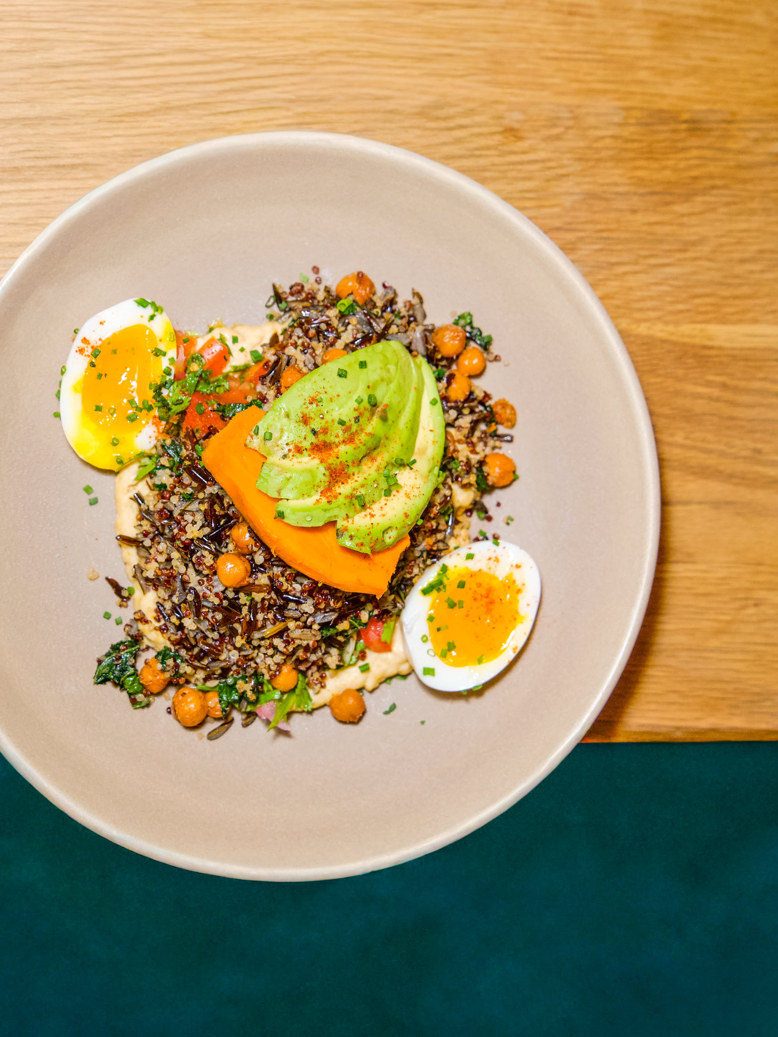 A bowl of mixed grains salad garnished with chopped chives, topped with half a boiled egg, a slice of avocado sprinkled with paprika, and a slice of tomato, on a wooden surface.