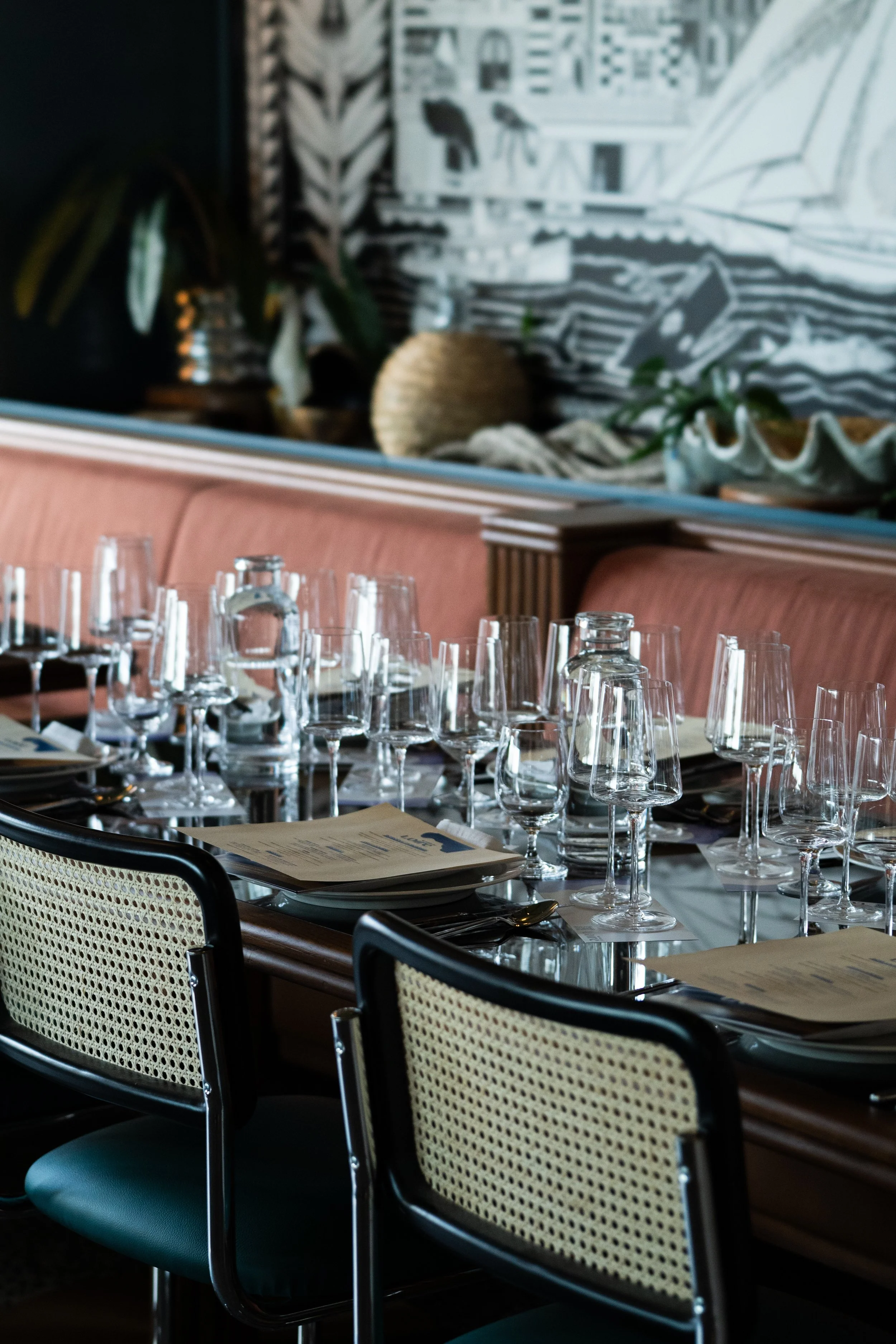 Indoor dining setting with tables set with glassware, utensils, and menus. Chairs are upholstered with patterned fabric, and the background features a mural and decorative plants.