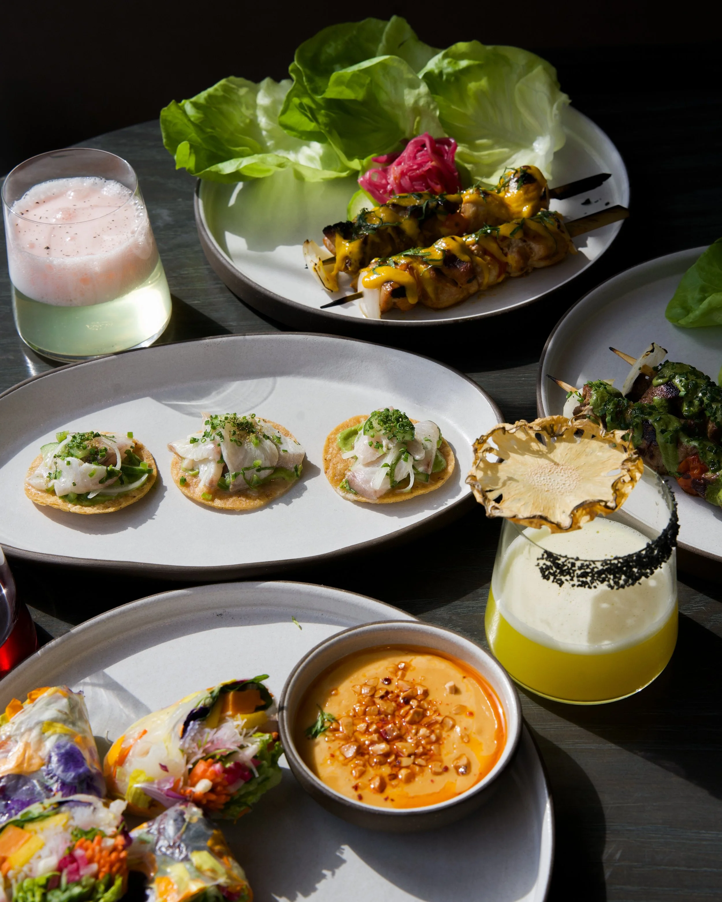 A variety of colorful dishes on a black table, including grilled chicken skewers with yellow sauce, small open-faced appetizers with seafood and greens, a bowl of creamy orange soup with nuts, and a beverage with a pineapple slice and salted rim, gar