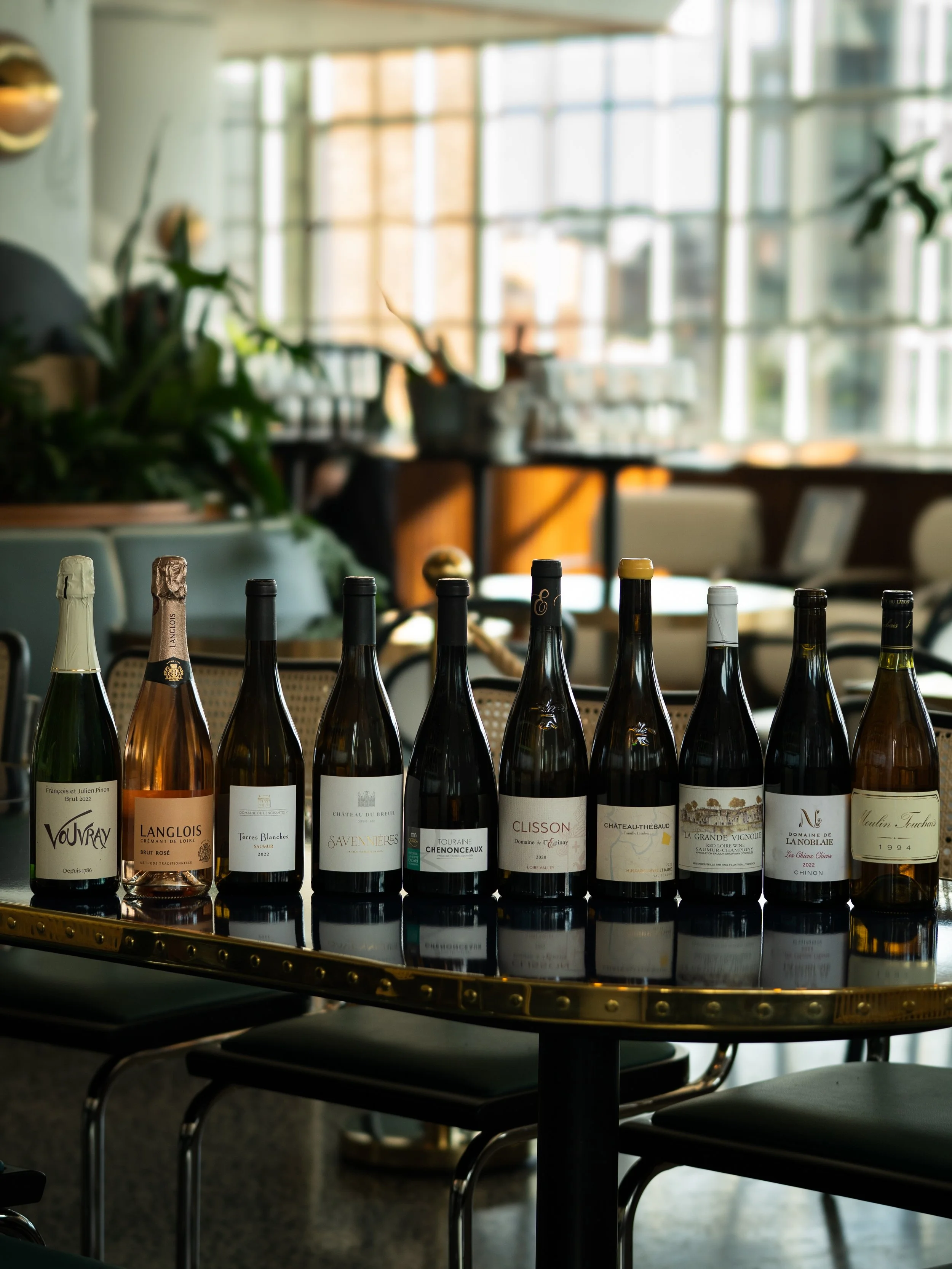 A row of wine bottles on a reflective black surface with a blurry restaurant or cafe interior in the background, featuring large windows and potted plants.
