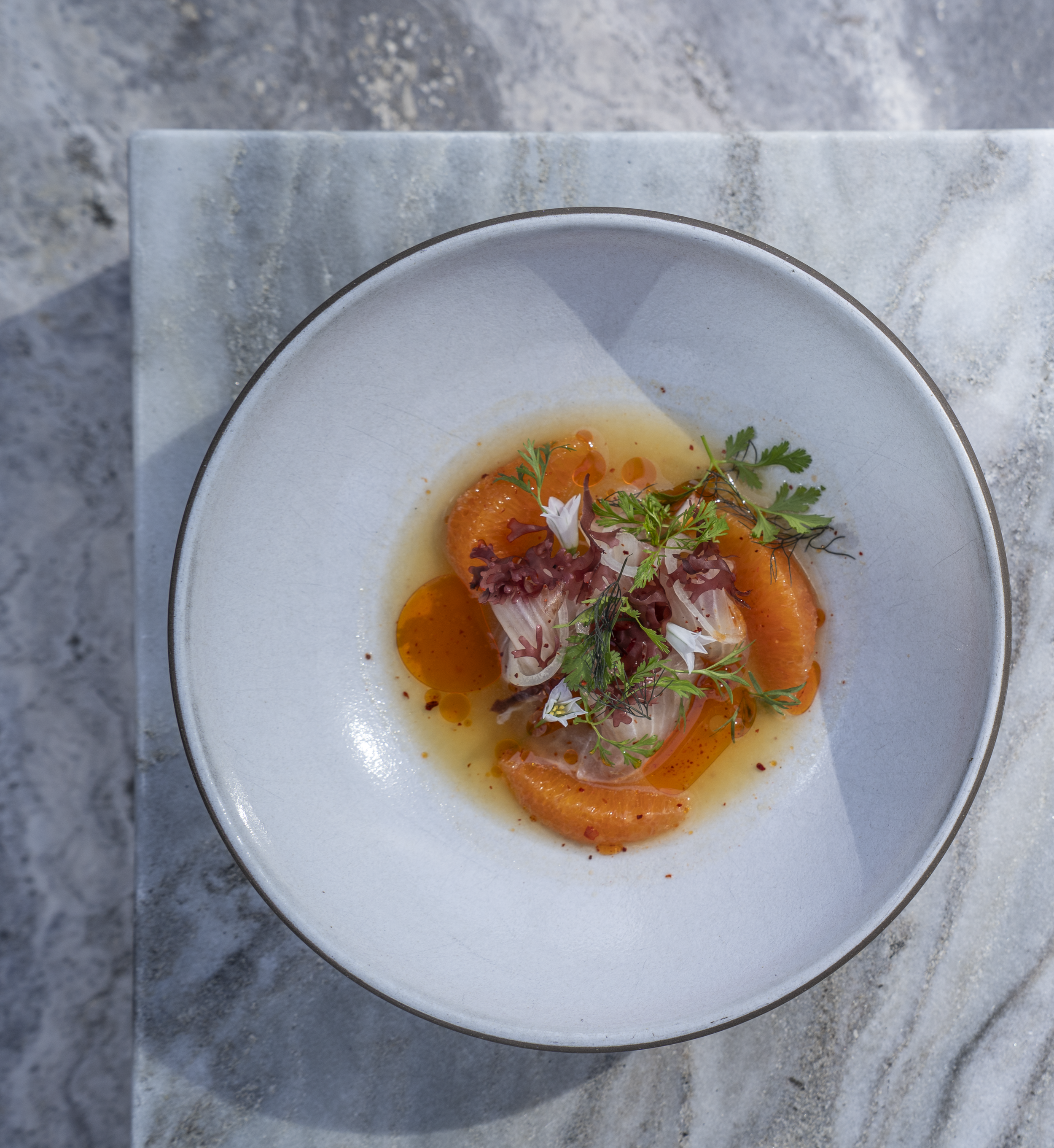Overhead view of a white bowl containing a poached egg garnished with herbs and red chili flakes, placed on a stone surface.