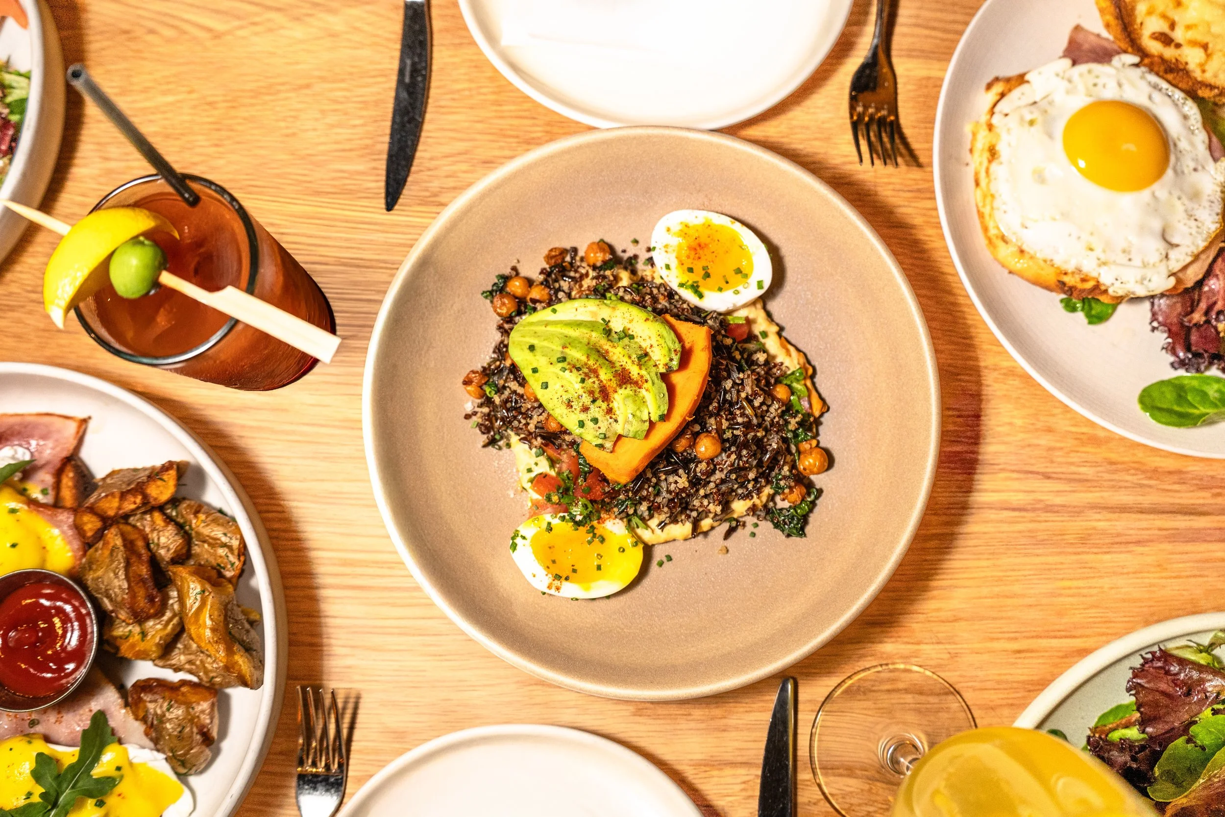 Plate of cooked quinoa salad topped with sliced avocado, halved boiled eggs, and chopped herbs, surrounded by glasses and other dishes on a wooden table.