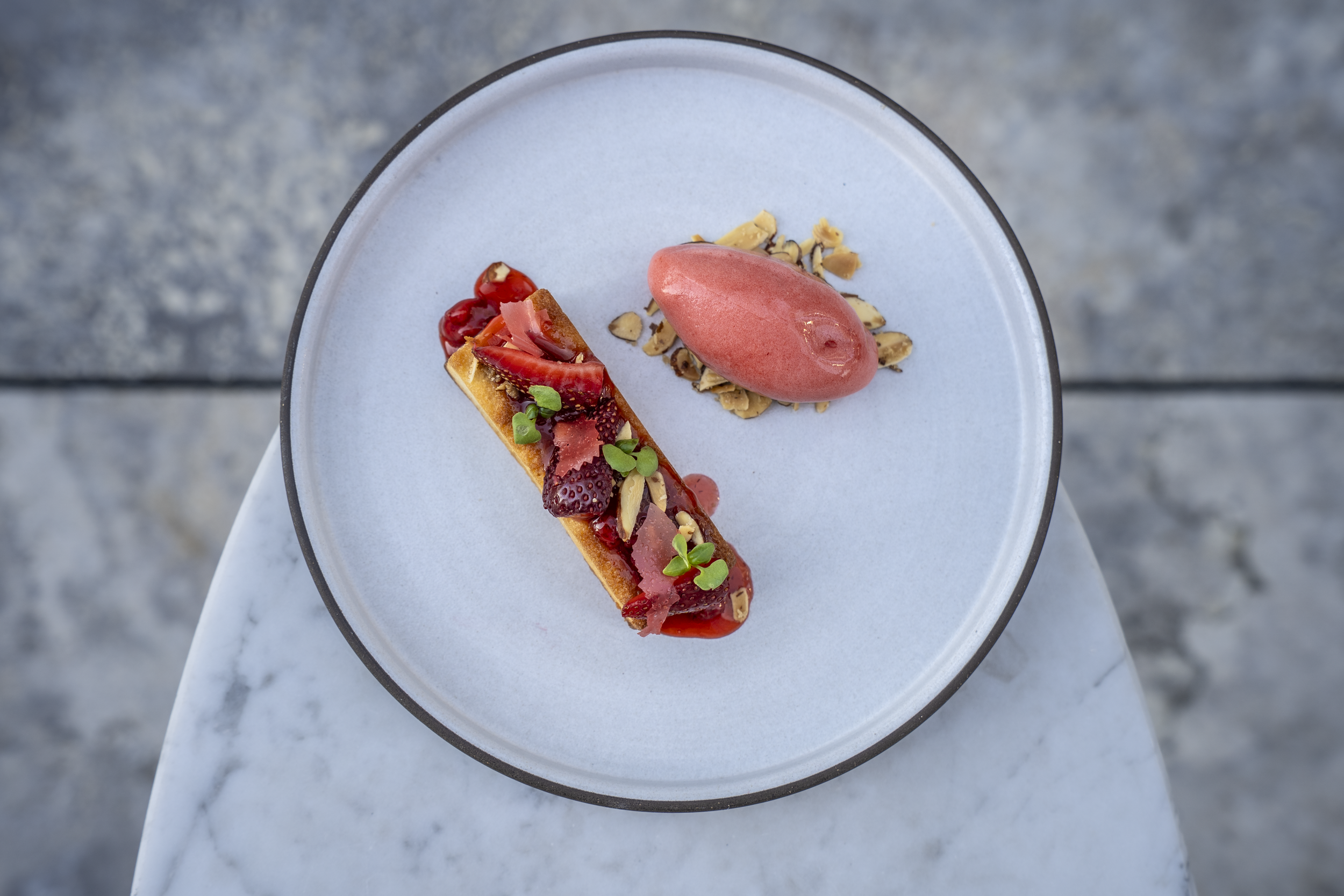A dessert plate with a rectangular tart topped with sliced strawberries, pomegranate seeds, and green garnish, alongside a pink sorbet and chopped nuts.