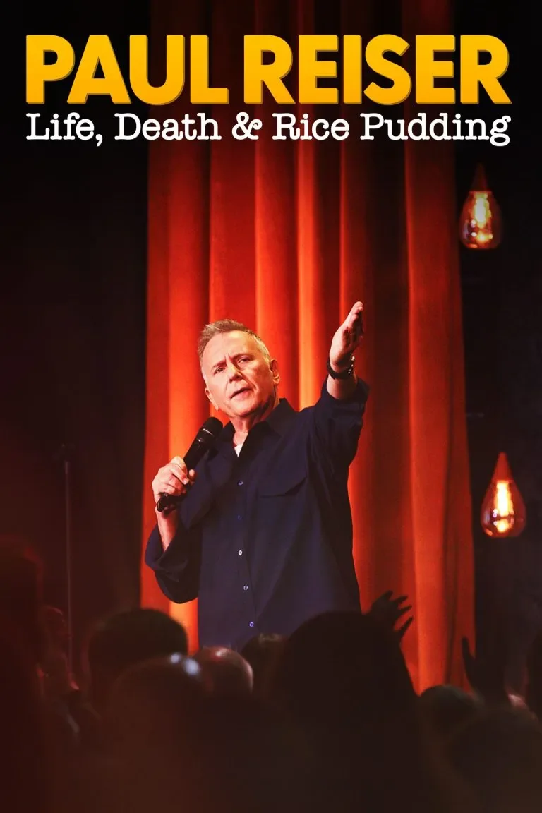Paul Reiser performing stand-up comedy on stage with orange curtains, holding a microphone in his right hand, and gesturing with his left hand, in front of an audience. "Paul Reiser: Life, Death and Rice Pudding"