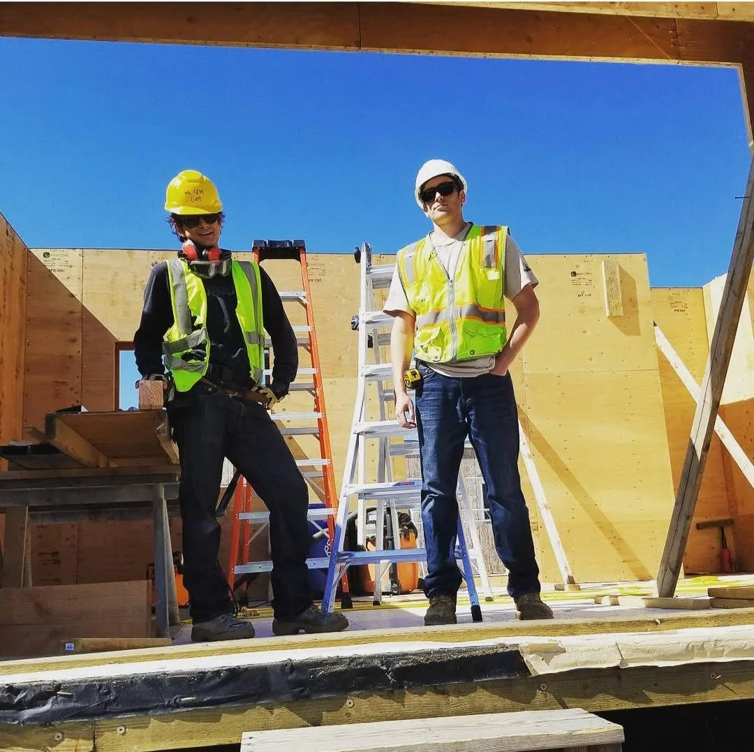 Two construction workers standing on a building site, wearing safety vests and hard hats, with wooden walls and ladders behind them under a clear blue sky.