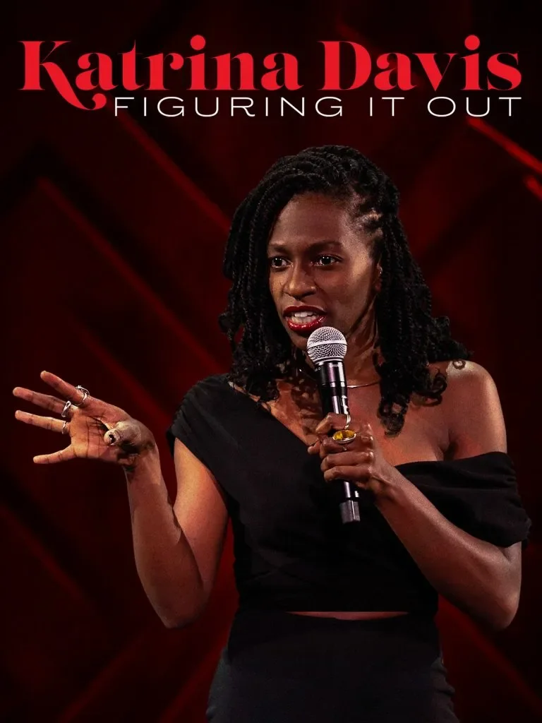 Katrina Davis performing stand-up comedy on stage with a microphone, wearing a black off-the-shoulder top, with the text 'Katrina Davis Figuring It Out' in red and white above her.