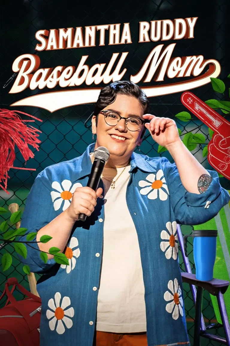 A person holding a microphone, wearing glasses, smiling, and adjusting glasses with one hand. They are wearing a denim shirt with large daisies and standing in front of a backdrop that reads 'Samantha Ruddy Baseball Mom' with decorative elements including a red foam finger, green leaves, and a ladder with a water bottle.