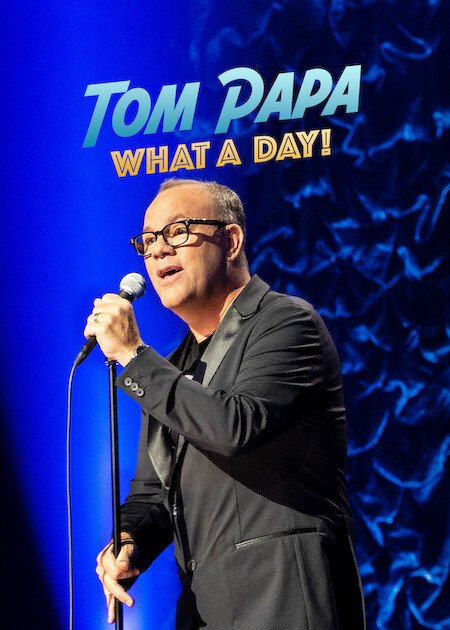 A man wearing glasses and a black suit singing into a microphone on stage with a blue background featuring the text 'TOM PAPA: WHAT A DAY!'.