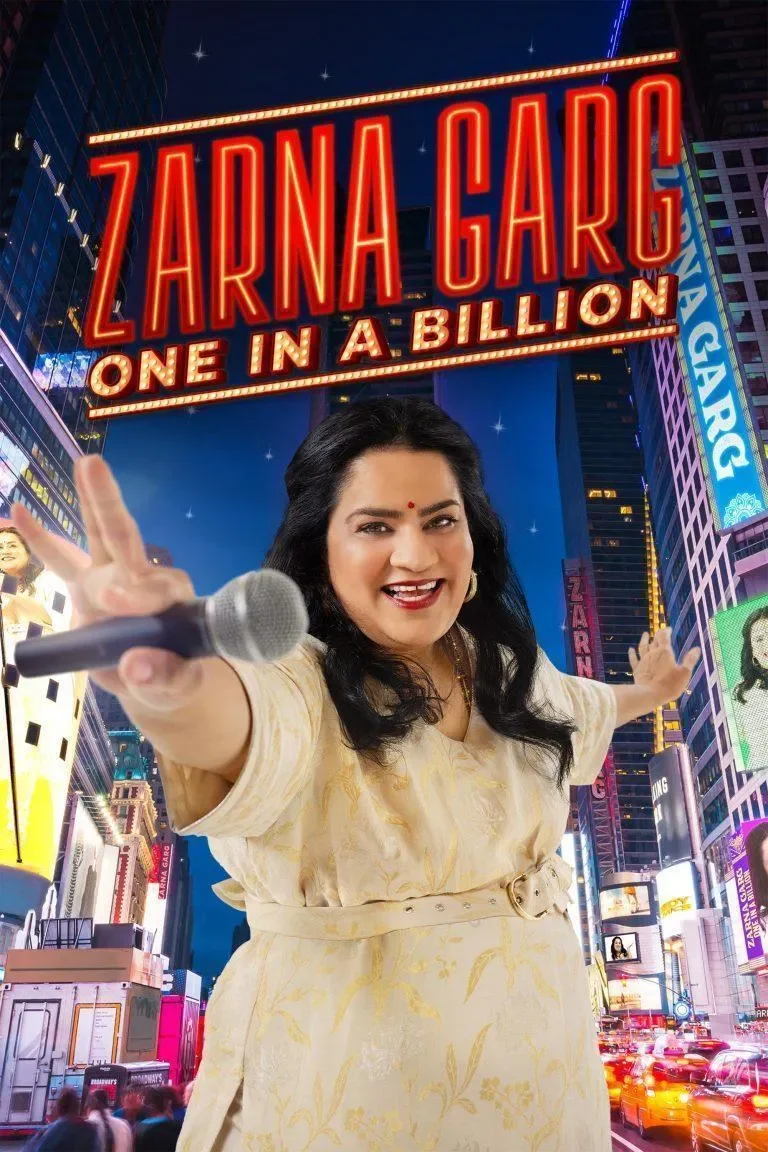 A woman in a cream-colored dress standing in front of a Times Square like cityscape at night, with a large neon sign that reads 'Zarna Garg One in a Billion,' holding a microphone and making a peace sign with her hand.