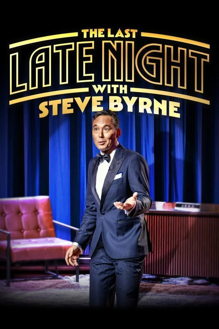 Comedian Steve Byrne performing on stage for "The Last Late Night with Steve Byrne" show. He is wearing a dark suit and bow tie, gesturing with one hand, with a blue curtain background and a pink armchair to the side.
