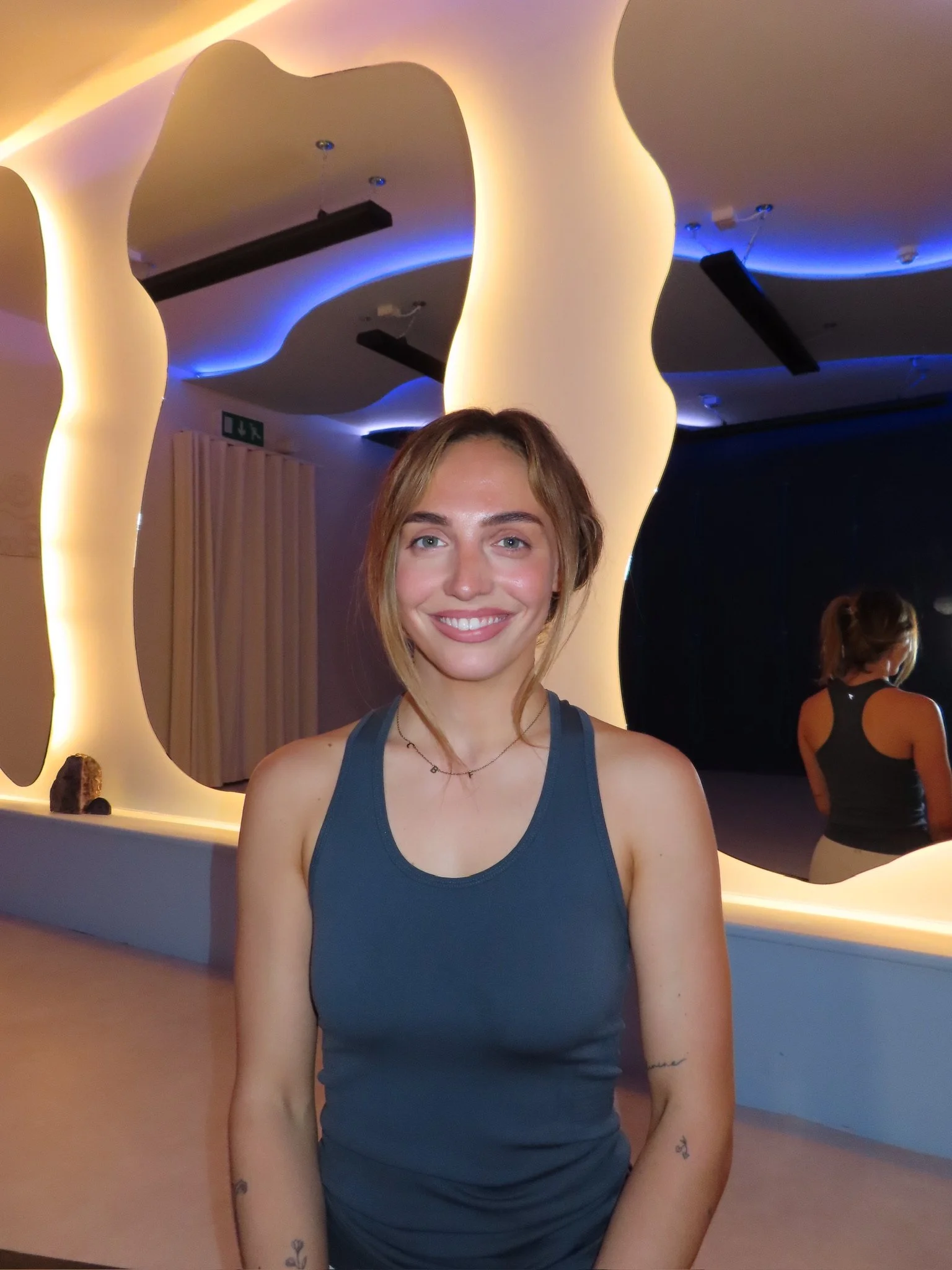 A smiling woman with shoulder-length light brown hair and tattoos on her arms, wearing a blue workout top, standing in front of an artistic mirror with wavy outlines and soft lighting.