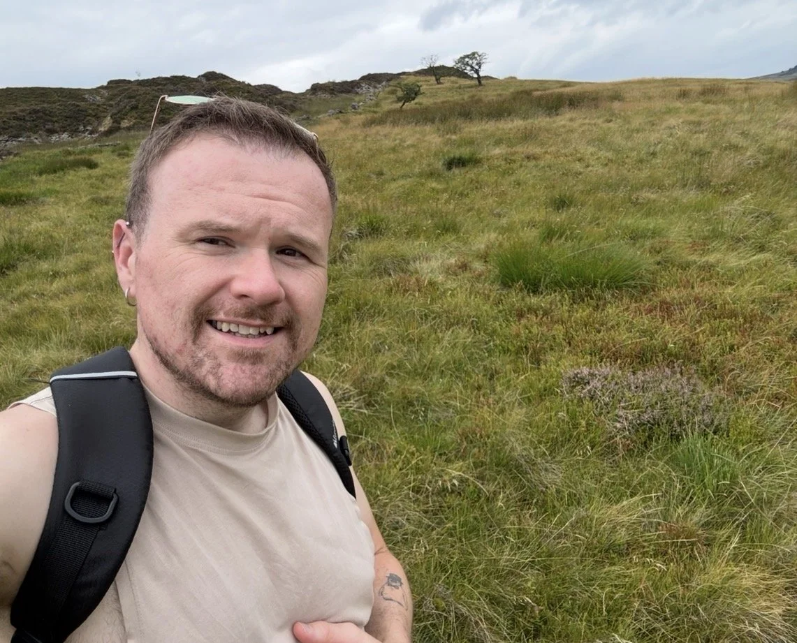 A man smiling in a grassy outdoor landscape with rolling hills and sparse trees in the background.