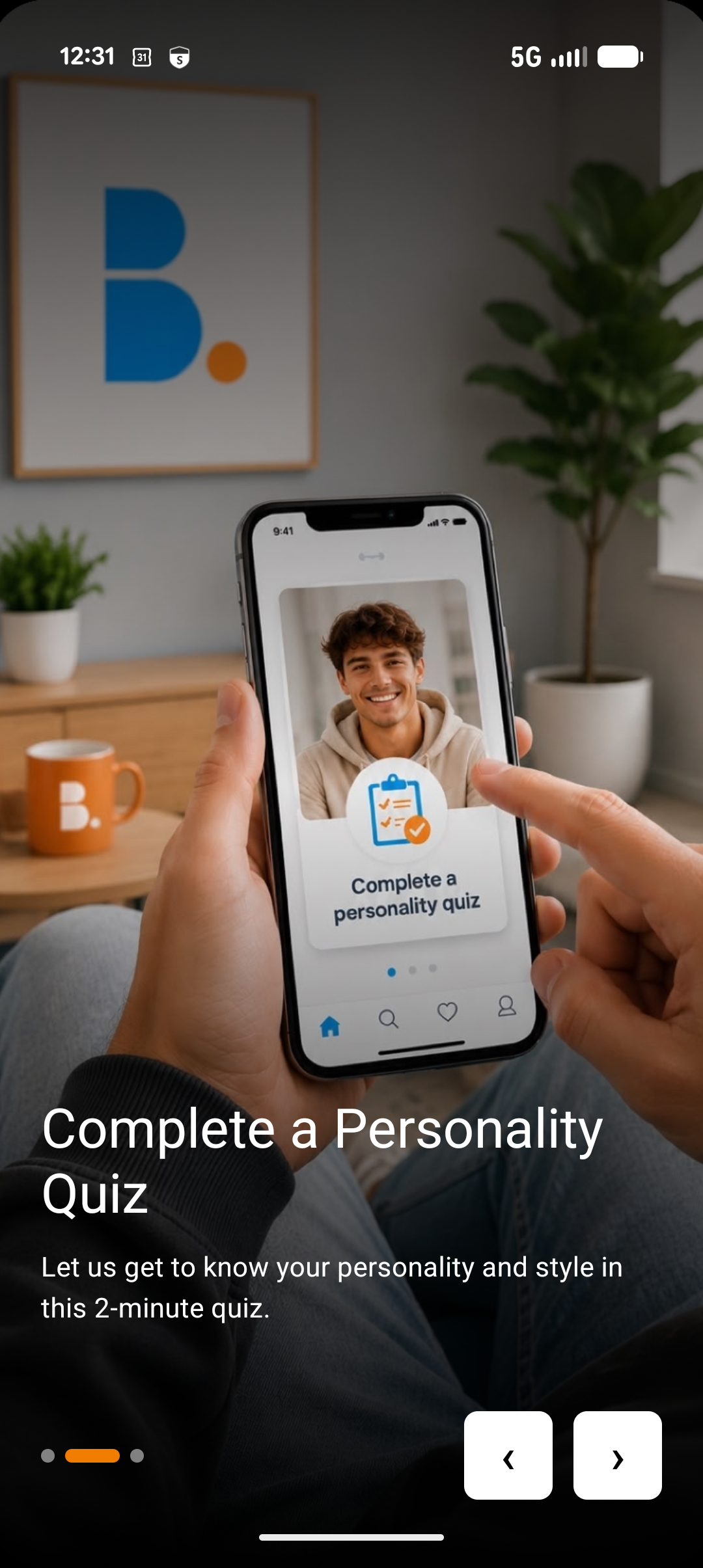 A person holding a smartphone with a screen displaying a man smiling and an app prompt to complete a personality quiz. The background shows a living room with a framed abstract art piece, a potted plant, an orange mug with a white logo, and a wooden table.