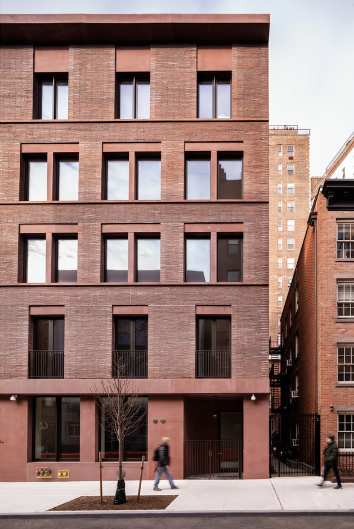 A modern red-brick apartment building with large windows and a sidewalk in front, with two people walking by and a small tree.