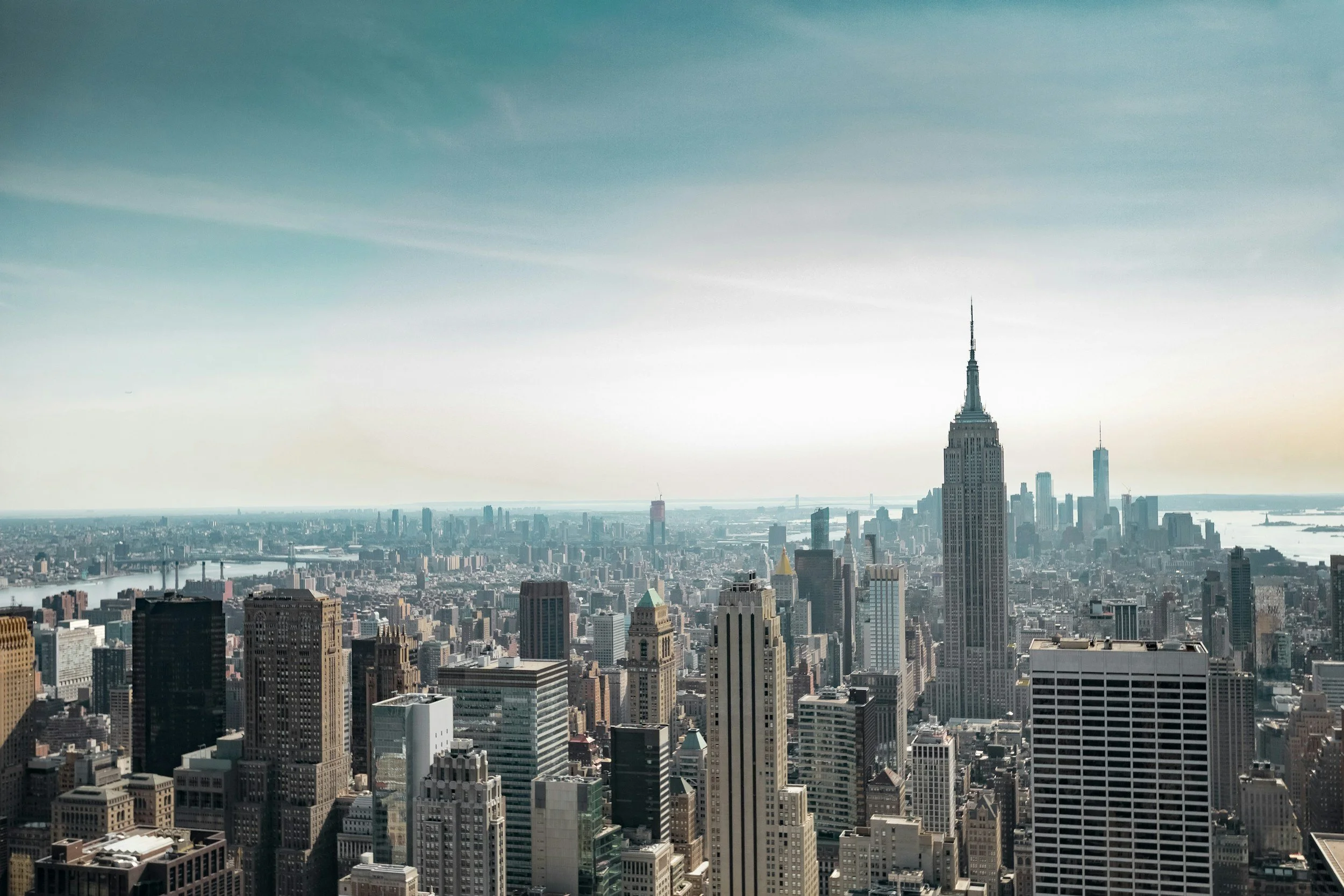 A cityscape view of Manhattan, New York City, featuring the Empire State Building tall among other skyscrapers with a river in the background and a clear sky.