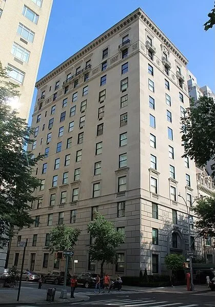 A tall, beige, multi-story building with many windows, located on a city street with trees and pedestrians in the foreground.