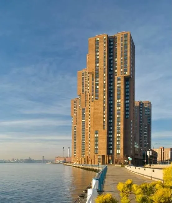 A tall, modern high-rise building with a stepped design, located by a body of water, with a walkway and some yellow plants in the foreground, under a partly cloudy sky.