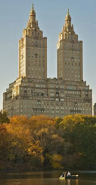 Two tall, light-colored skyscrapers with spire-like tops rising above trees in a park, with a small boat on a water body in the foreground.