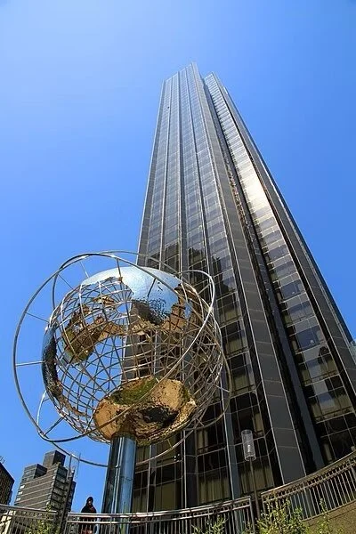 A tall modern skyscraper with reflective glass windows and an art installation of a metallic globe with an inscribed map of the world on a pole in front.