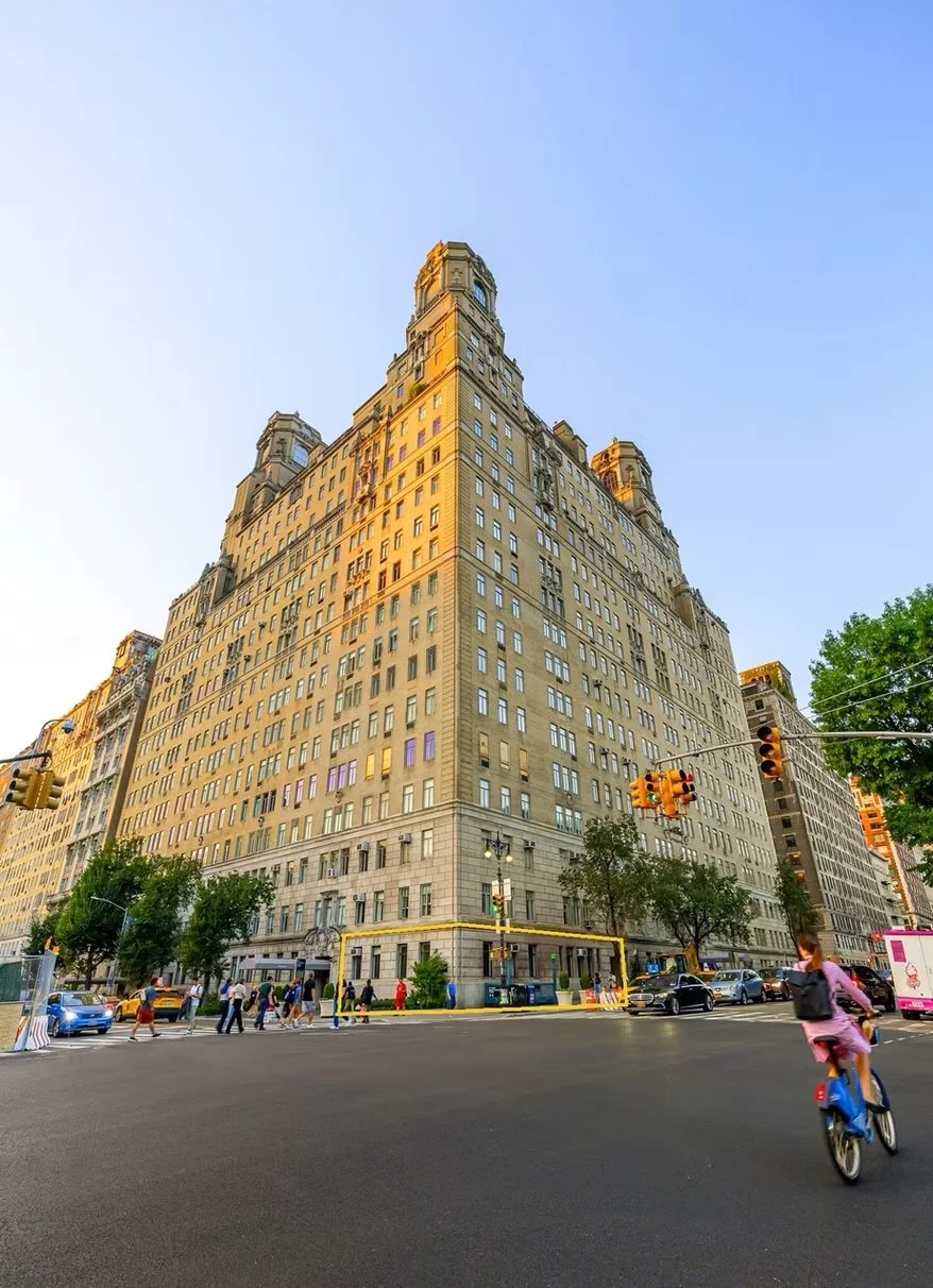 Tall beige building on a city street with traffic lights, people walking, a person riding a bicycle, and trees in the foreground during daytime.