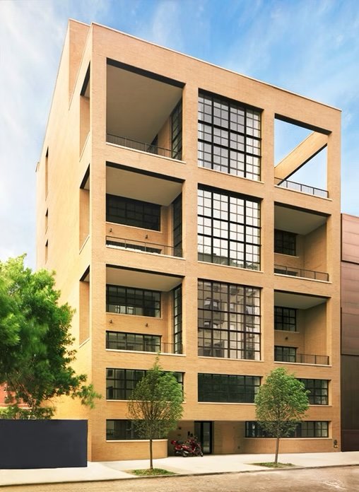 Modern beige apartment building with large black-framed windows and open balconies on each floor, surrounded by small trees and a sidewalk.