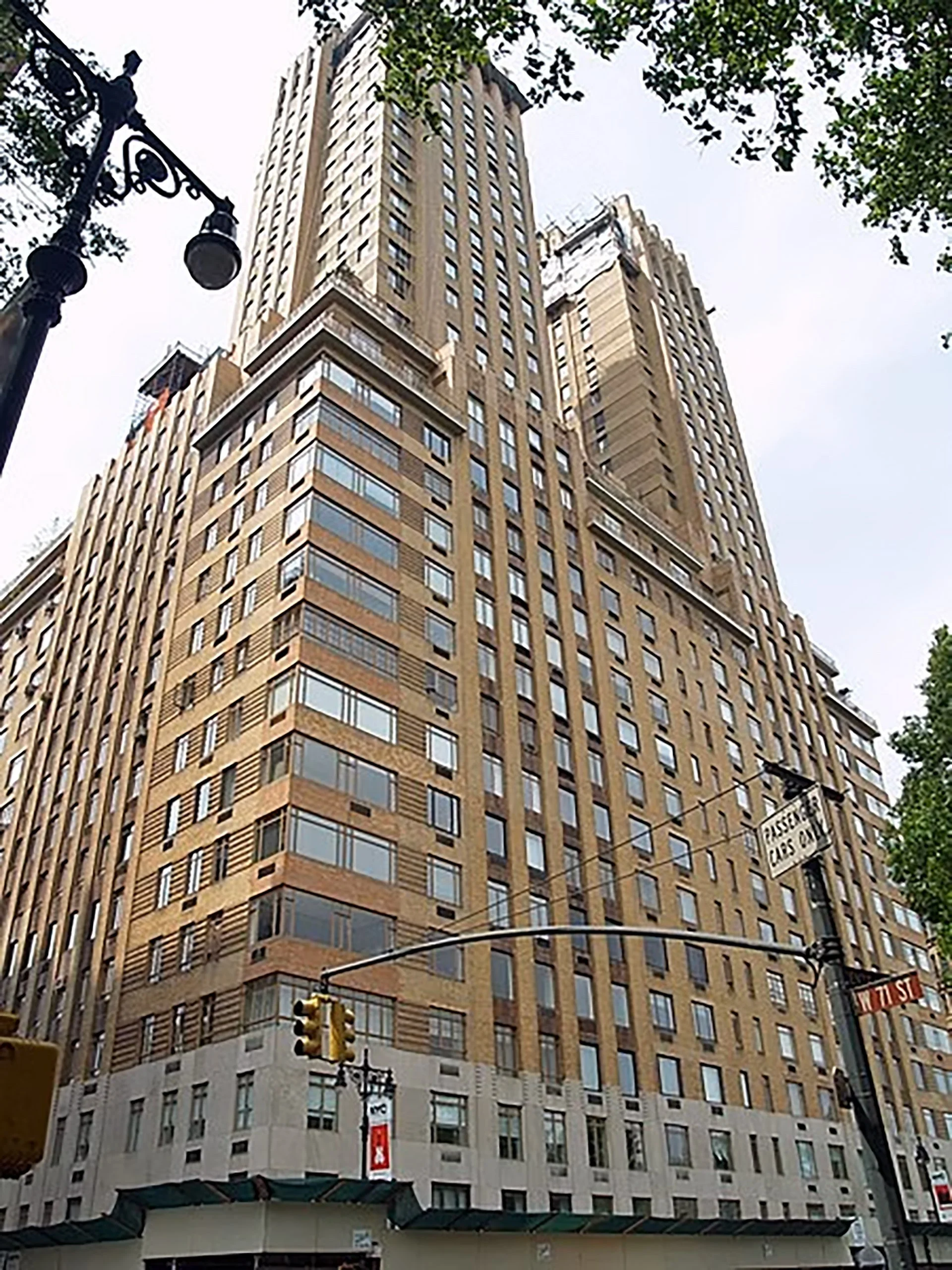 Tall beige and brown skyscraper with many windows, surrounded by street signs and a streetlamp, with trees and an overcast sky in the background.