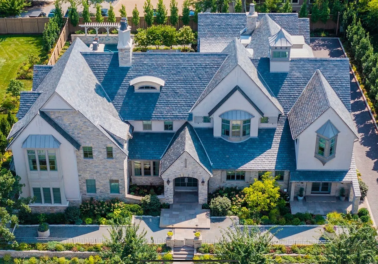 A large, multi-story house with a gray tiled roof, white and stone exterior walls, multiple gables and dormer windows, surrounded by green landscaping and a backyard with a pool and seating area.