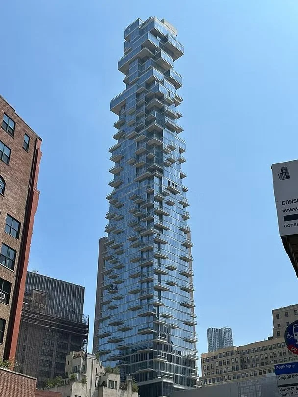 A tall, modern skyscraper with an irregular, stacked design featuring multiple glass balconies, set against a clear blue sky surrounded by older, traditional buildings.