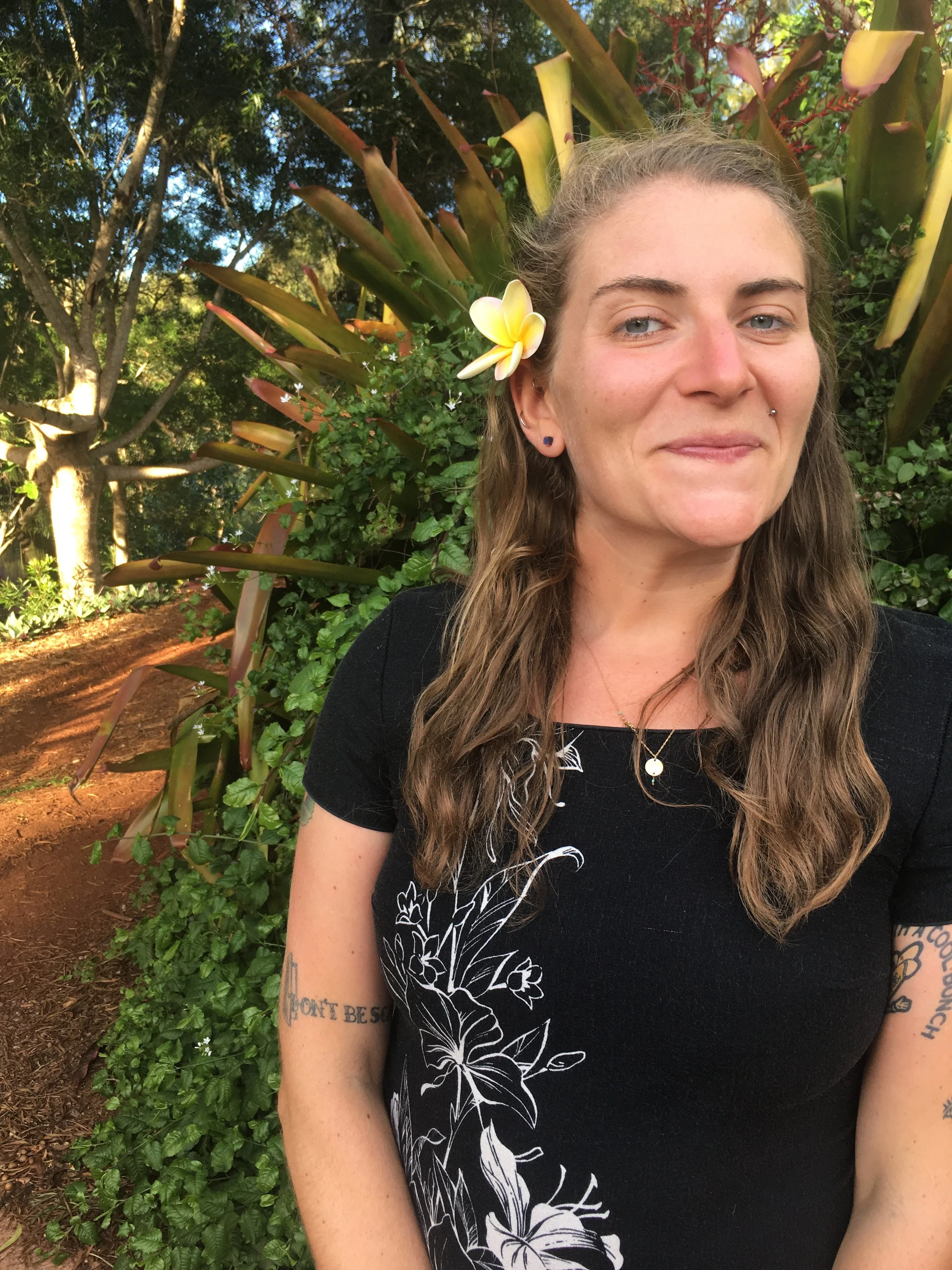 A woman with long wavy brown hair and tattoos on her arm, standing outdoors in a garden with lush greenery and flowering plants, wearing a black shirt with white floral designs and a yellow flower tucked behind her ear.