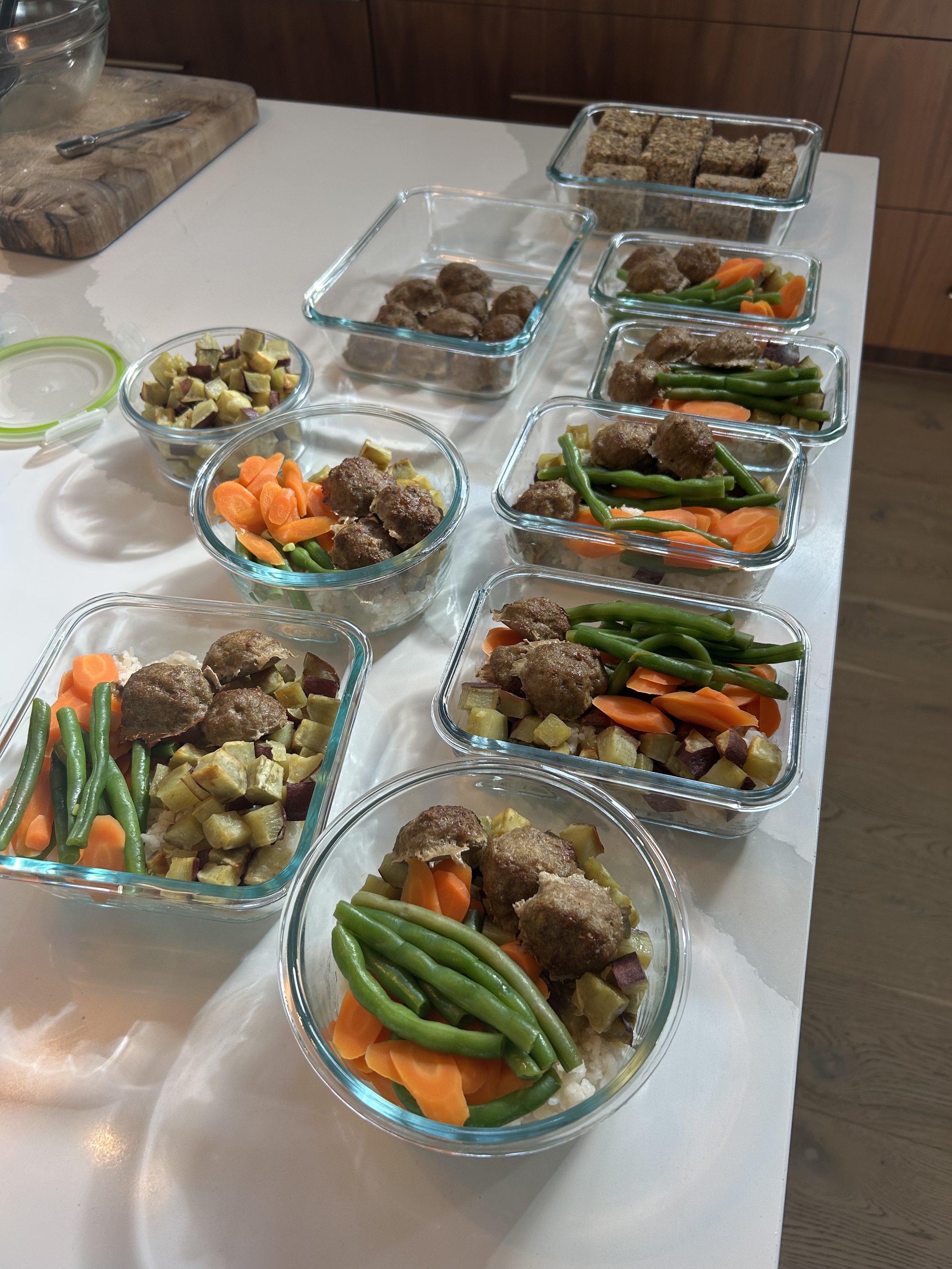 Glass containers with cooked vegetables and meatballs, including green beans, carrots, mashed potatoes, and veggie patties, arranged on a white kitchen counter.