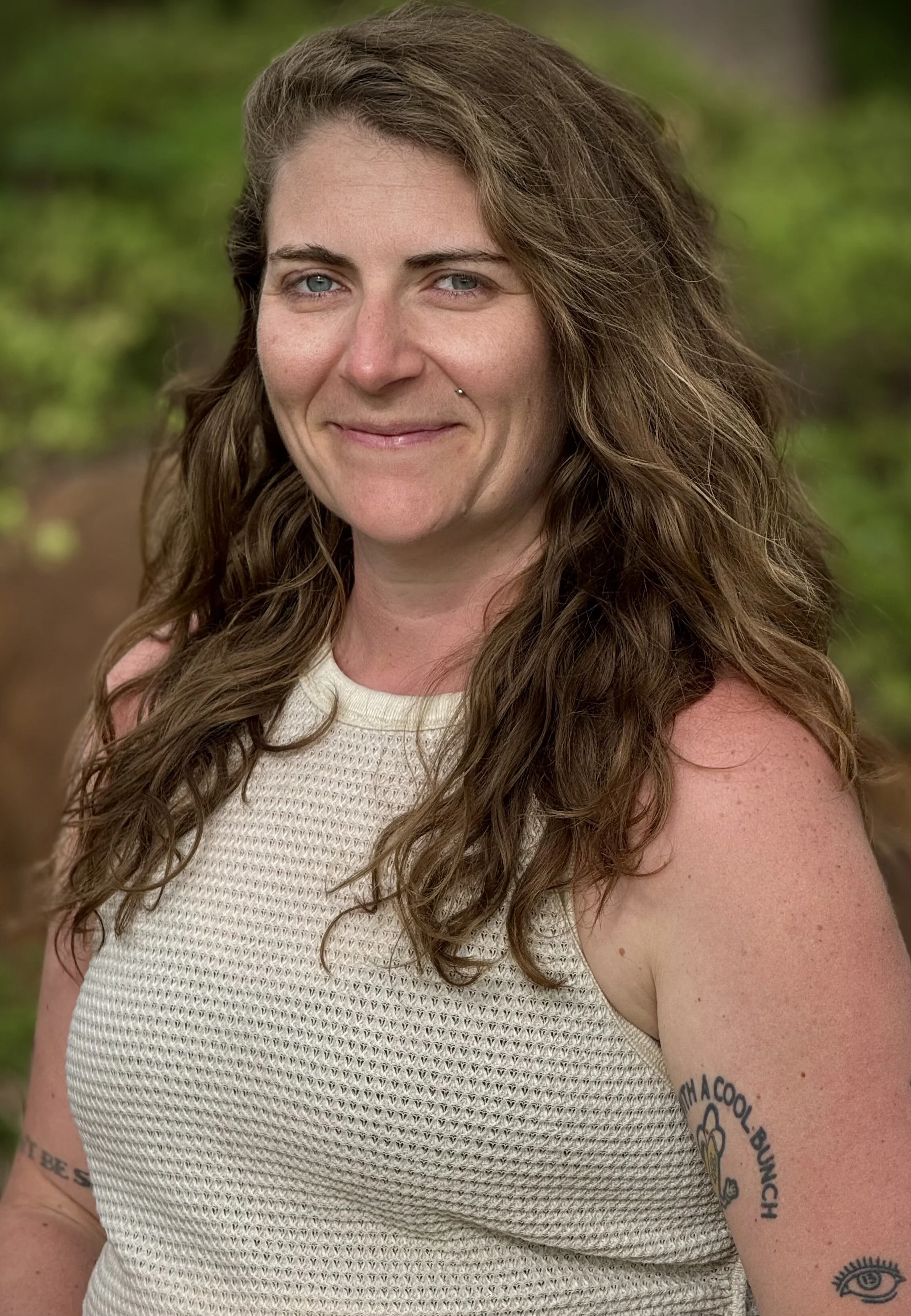 Close-up of a smiling woman with long wavy brown hair, blue eyes, and tattoos on her arm, outdoors with green blurred background.