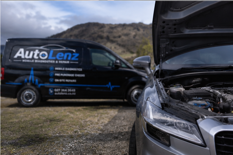 A silver car with the hood open, indicating engine trouble, parked on a grassy area with a hill in the background. A black mobile diagnostic and repair van labeled 'AutoLenz' is parked behind the car.