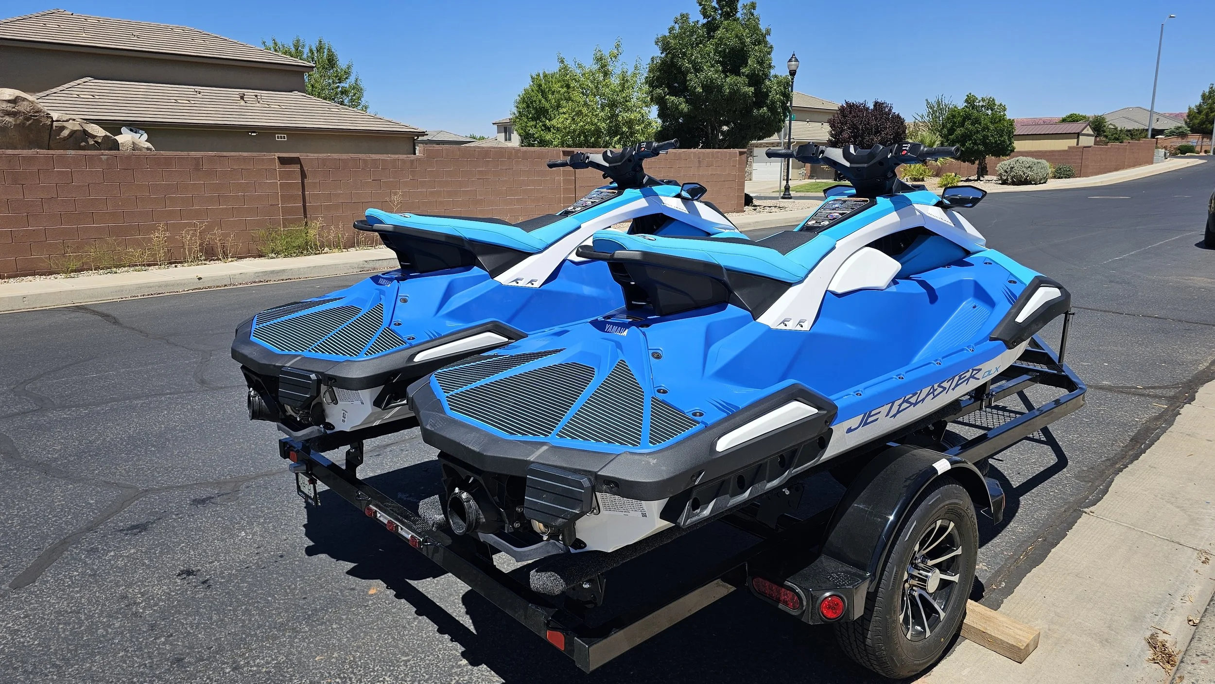 Two blue and white Yamaha JetBlaster FX personal watercraft on a trailer parked on a residential street with houses, trees, and a clear blue sky in the background.