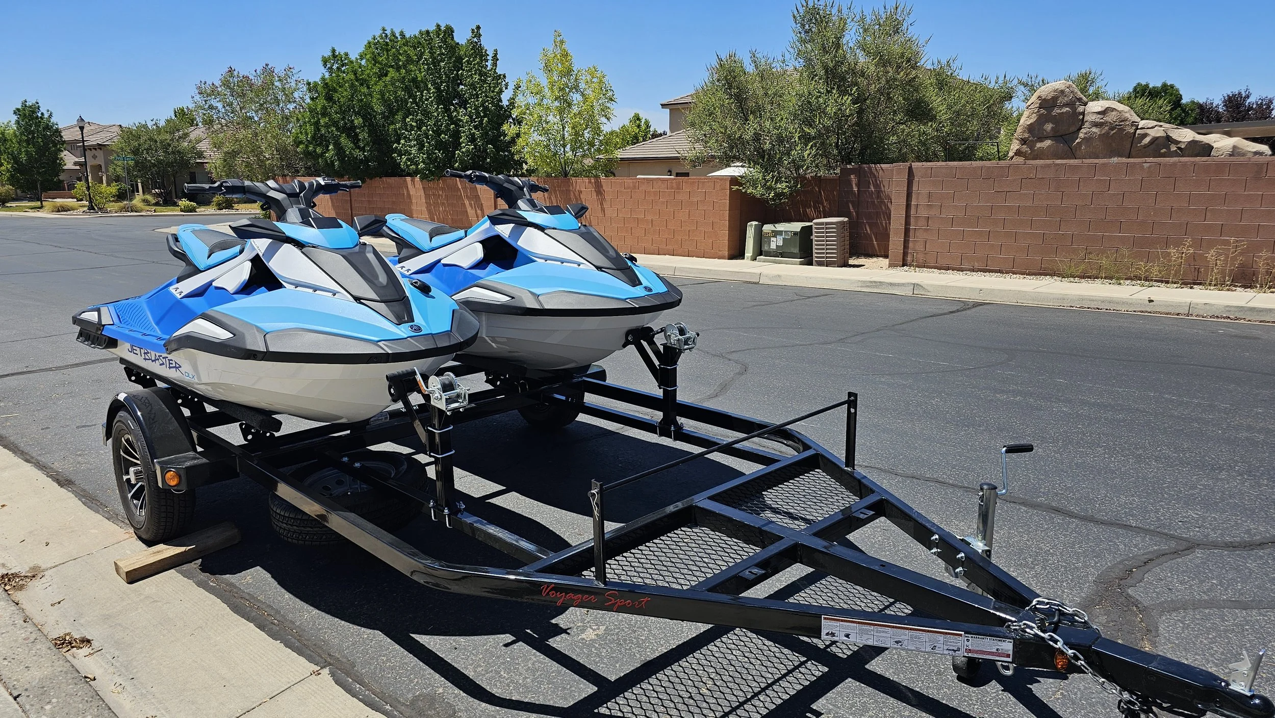 Two white and blue jet skis on a trailer on a residential street, with a brick wall and trees in the background.