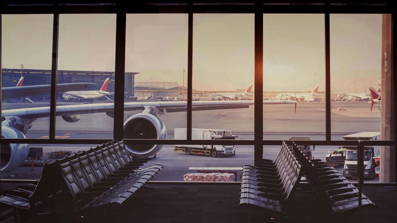 View of an airport terminal with empty black chairs facing large windows overlooking tarmac with parked airplanes, airport vehicles, and a sunset sky.