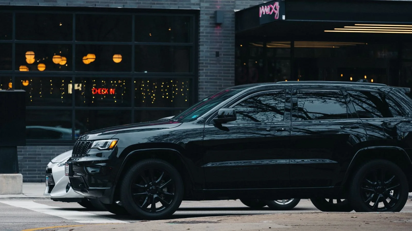 Black SUV and white car parked in front of a restaurant with large glass windows and warm yellow lights inside, with a neon sign reading 'Moxy' above the entrance.