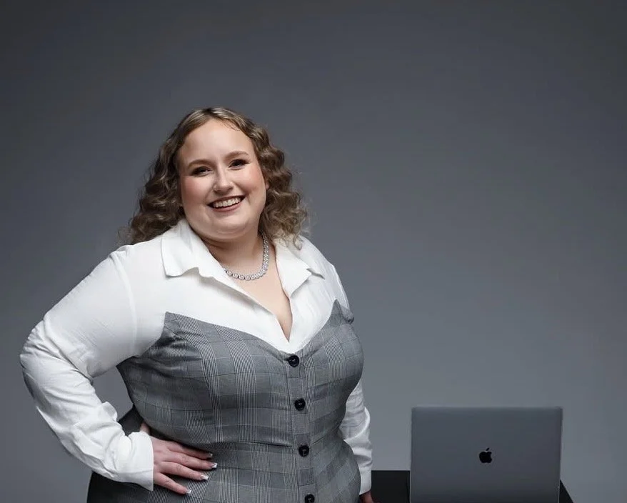 A woman with curly hair wearing a white shirt and gray vest, smiling, standing with hand on hip next to a laptop against a gray background.