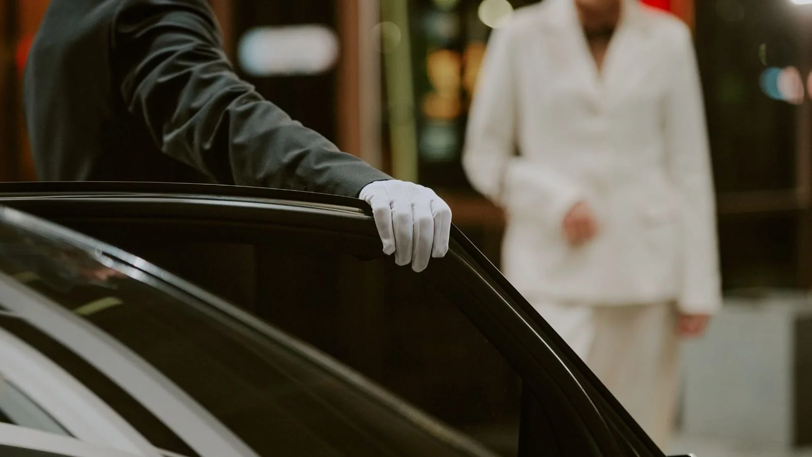 Person in dark clothing and white gloves opening or closing a car door, with a woman in white in the background.