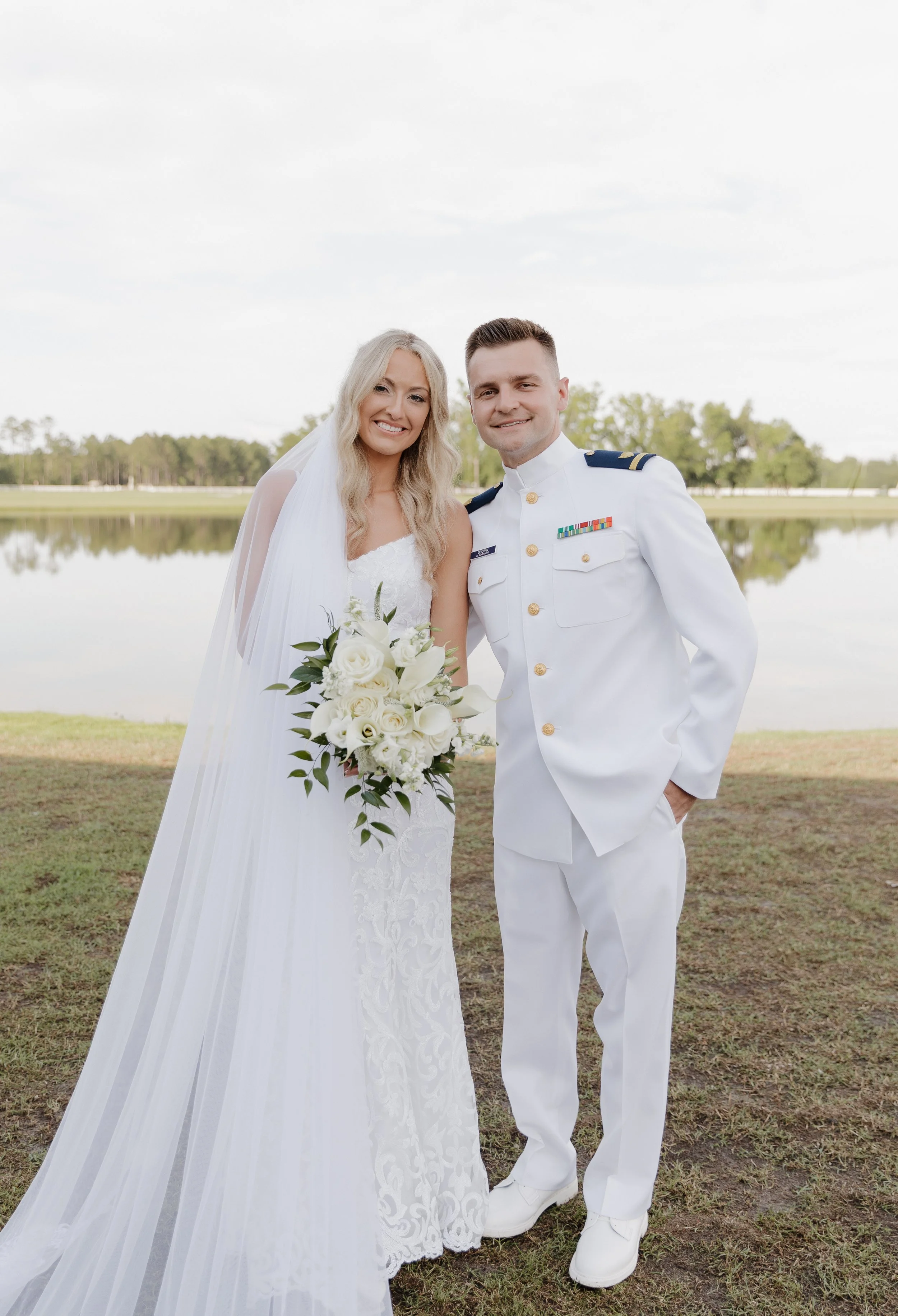 A bride and groom standing outdoors by a lake, smiling for a wedding photo, with the bride holding a bouquet of white flowers.