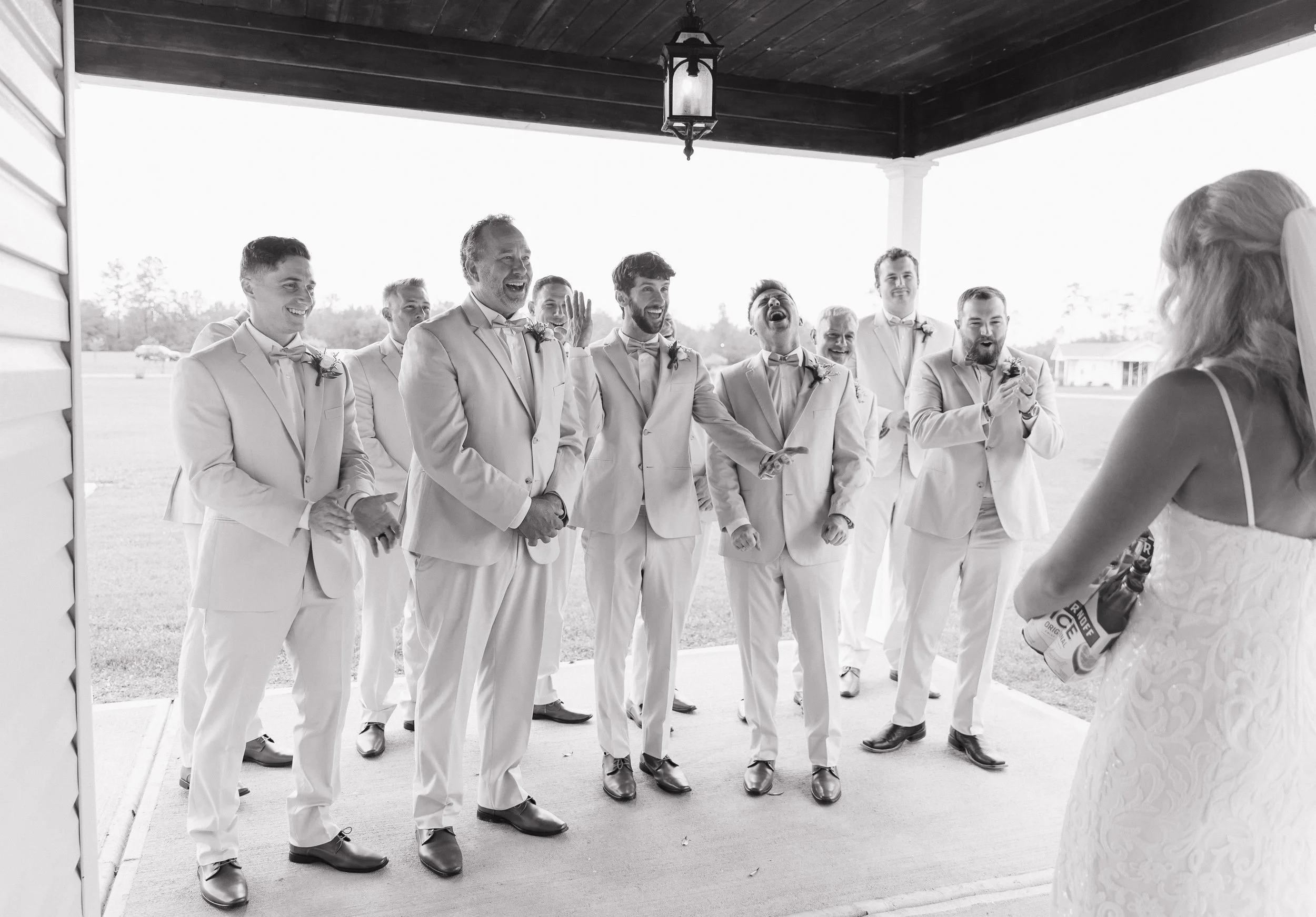 A group of men dressed in light-colored suits and bow ties are standing outdoors, laughing and smiling. A woman in a wedding dress is in the foreground facing the group. The setting appears to be a wedding celebration.