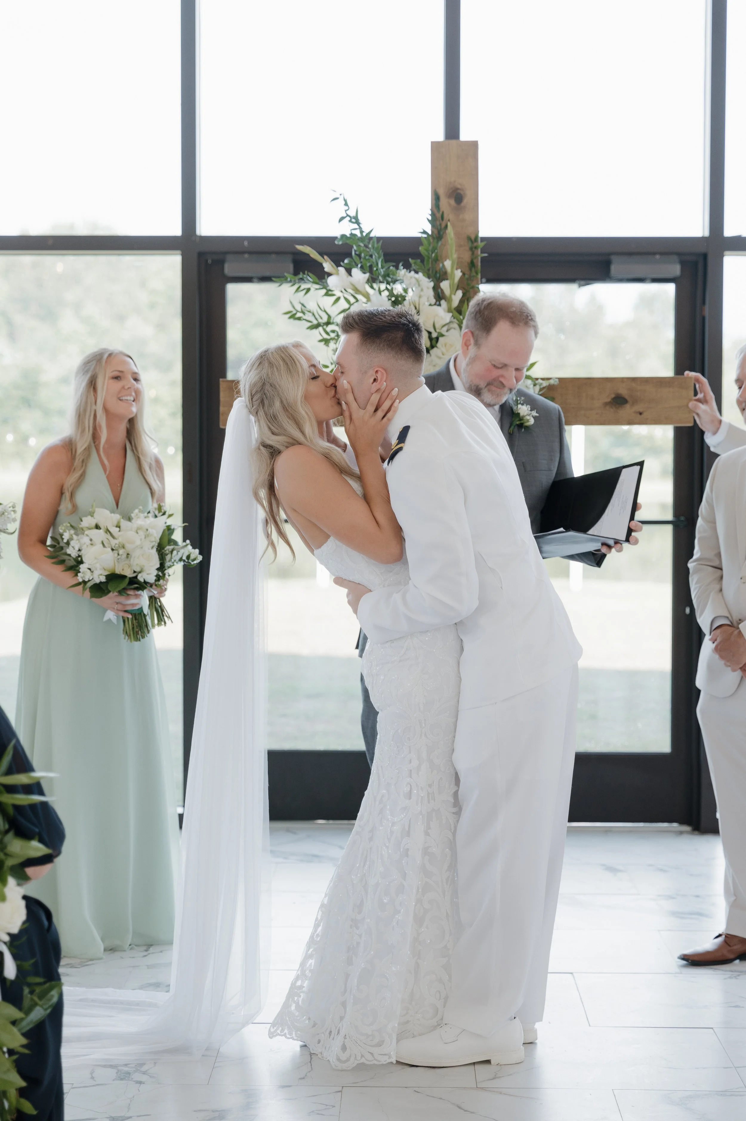 A bride and groom sharing a kiss during their wedding ceremony with a wedding party and a large floral cross in the background.