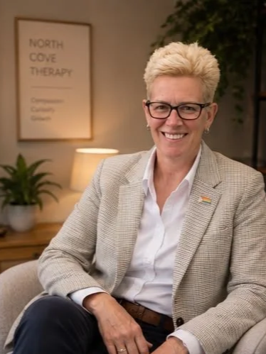 Smiling woman with short blonde hair and glasses sitting in an office or therapy room, wearing a light blazer and white shirt, with a framed poster and plants in the background.