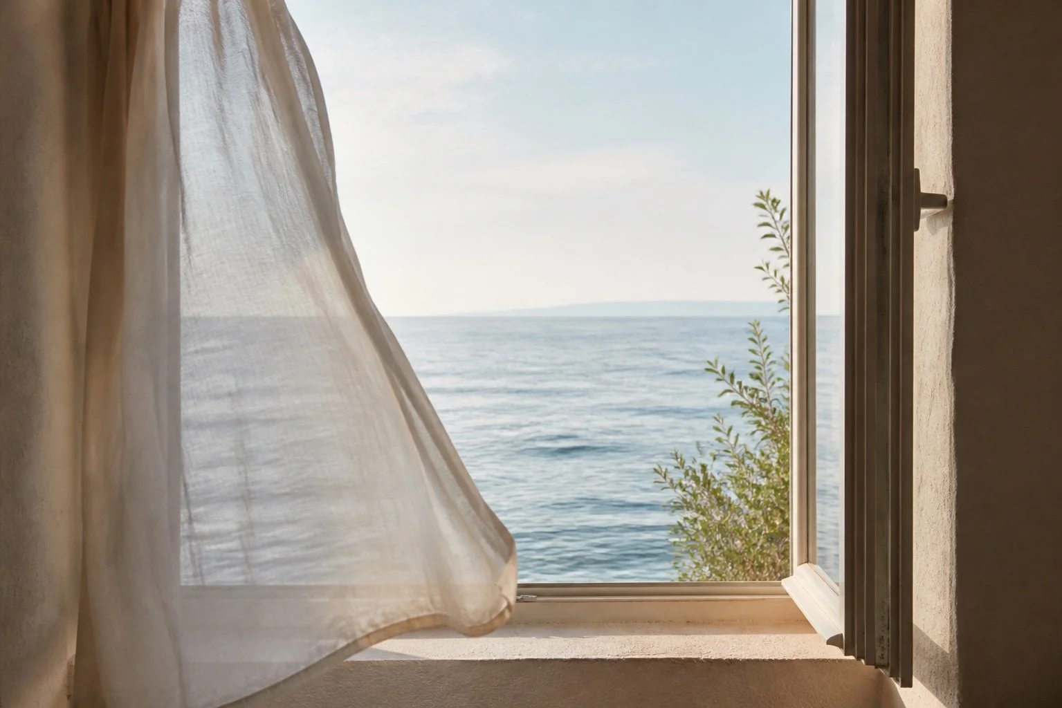 Open window with a white curtain blowing in the breeze, revealing a view of the ocean with a clear sky and some greenery outside.