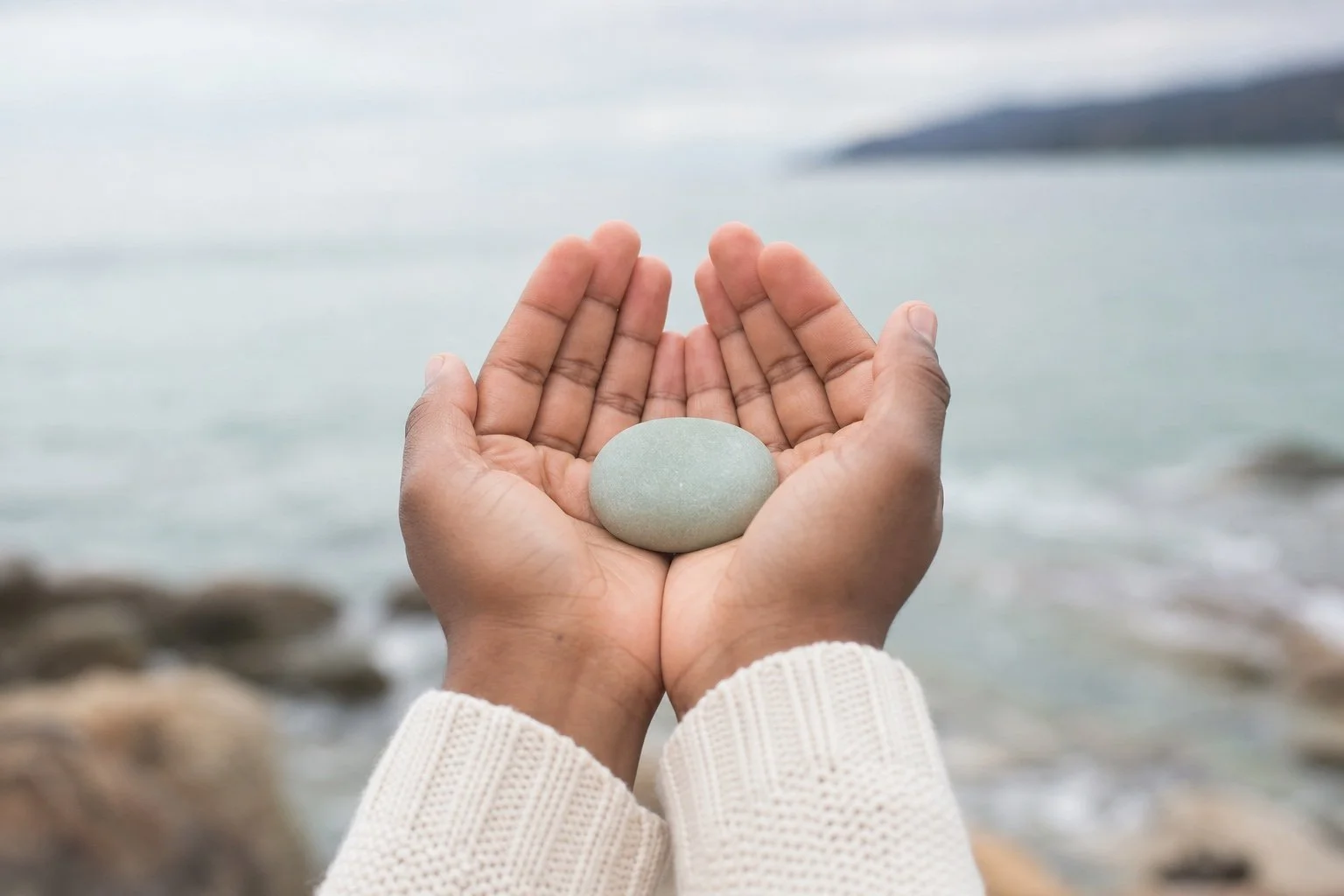 Two hands holding a smooth gray stone against a blurred ocean background.