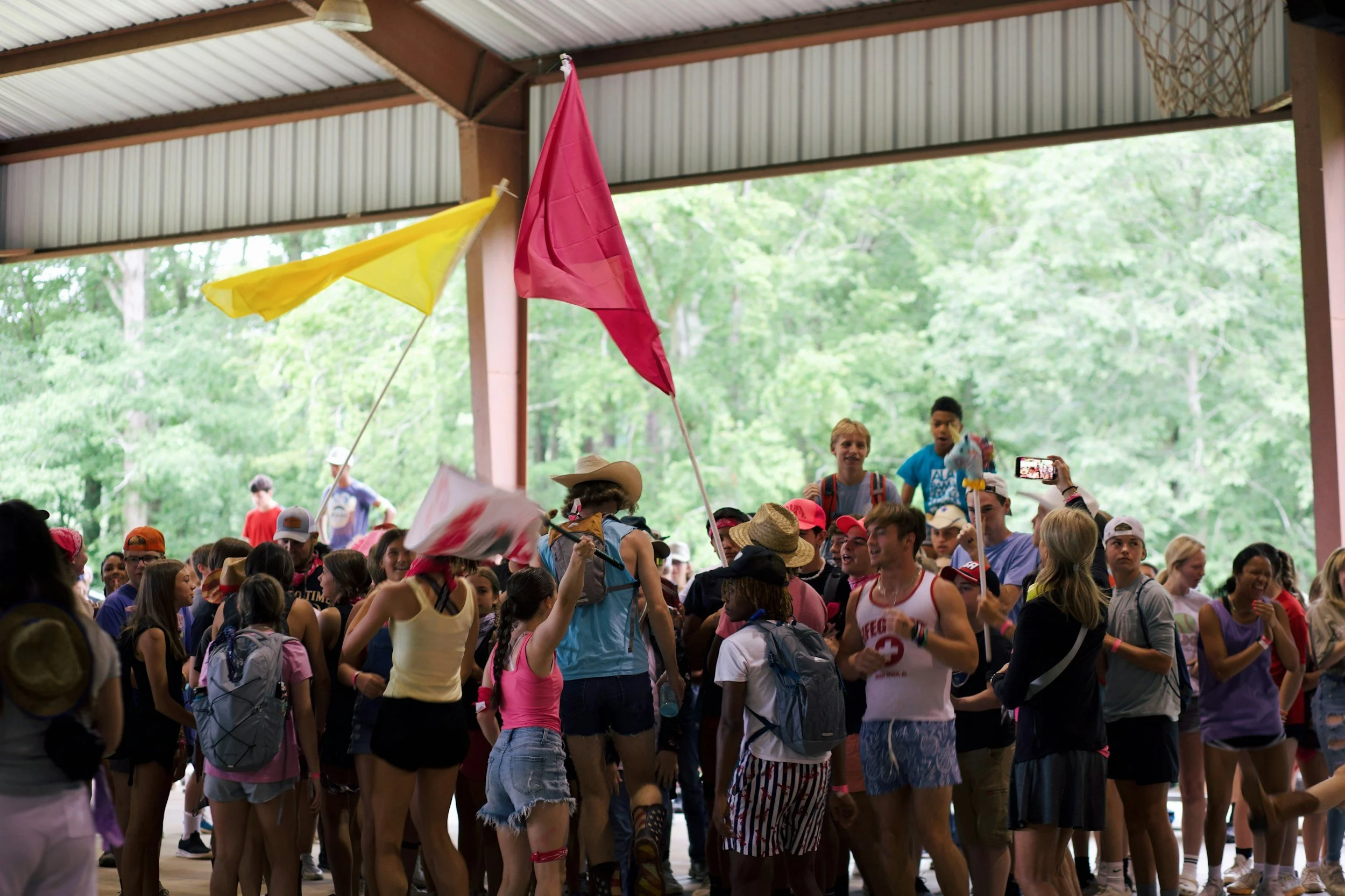 A large group of people gathered inside a covered pavilion, with some holding flags, wearing casual summer clothing, hats, and backpacks. The background features green trees visible through the open sides of the pavilion.