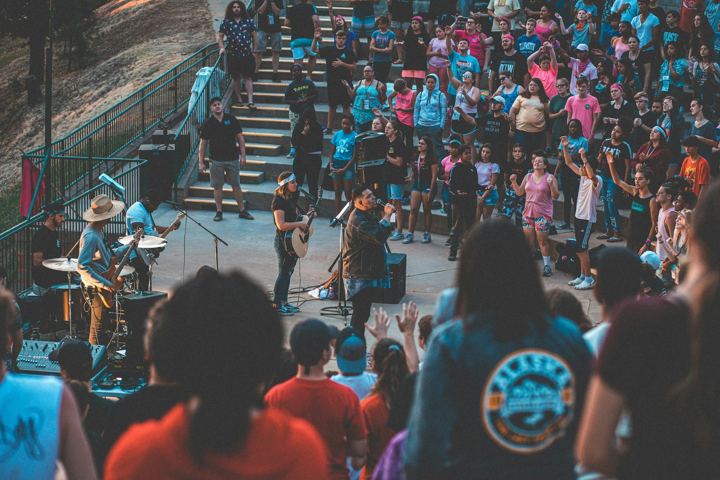 A large group of people, mainly children and teenagers, gathered outdoors on stone steps, participating in a singing or musical event. Some are raising their hands, while musicians at the front are playing guitars and singing into microphones. The surroundings include trees and rocky terrain.