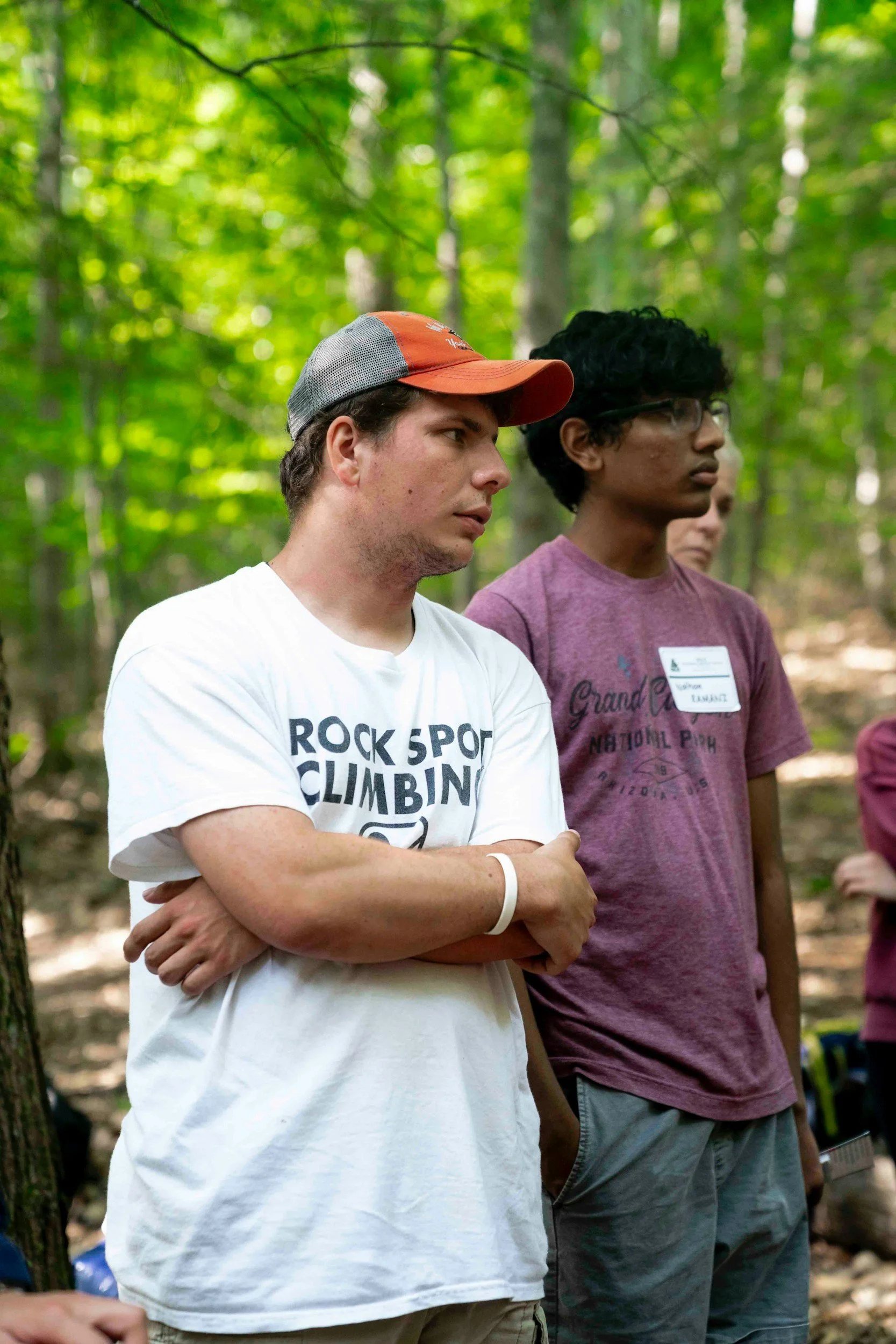 Two young men stand outdoors in a wooded area. The man in the white T-shirt has a baseball cap, while the man in the maroon T-shirt wears glasses. Both appear to be listening attentively.