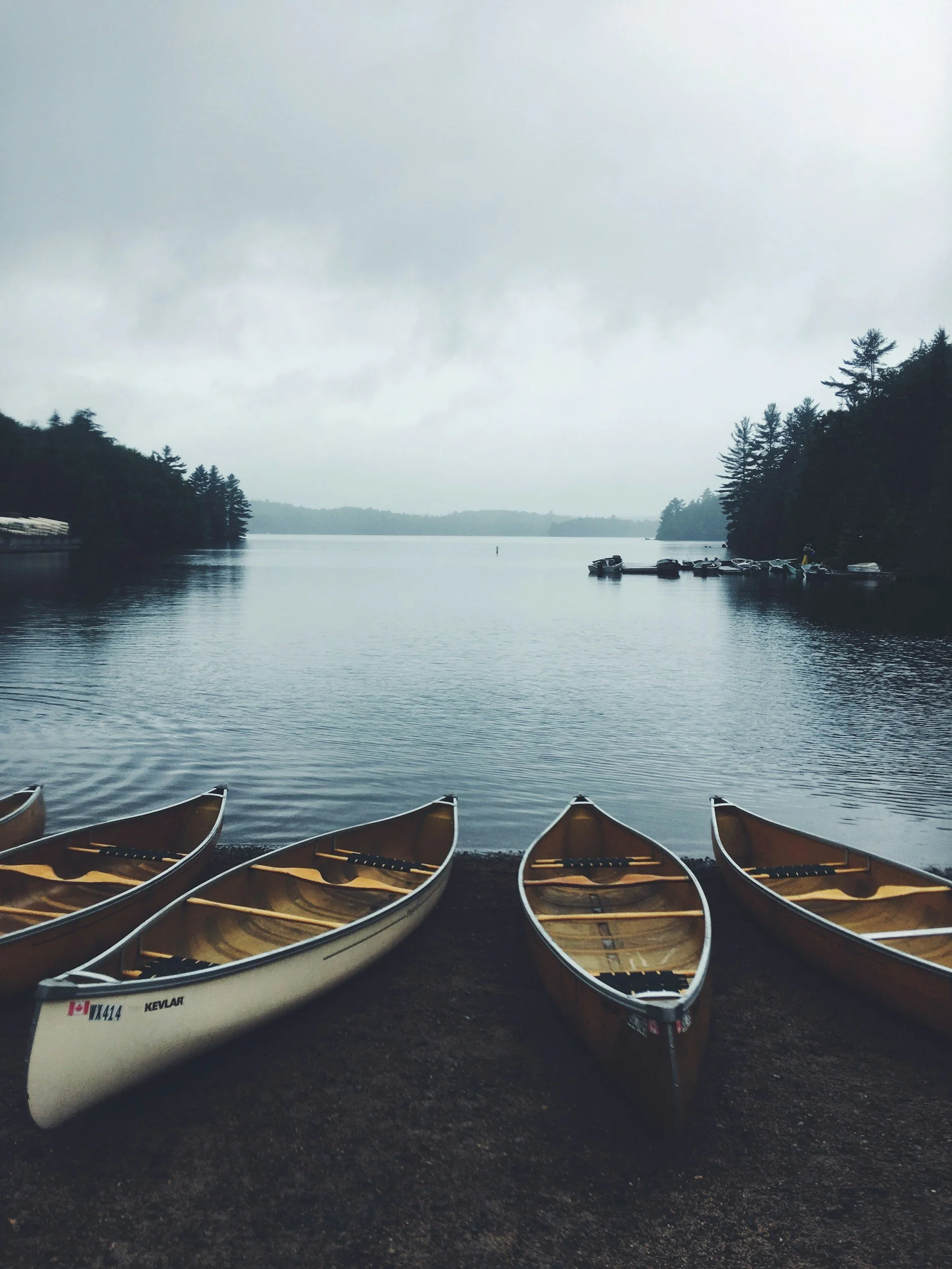 Multiple canoes are lined up on a shore facing a calm lake with trees on both sides and overcast skies.