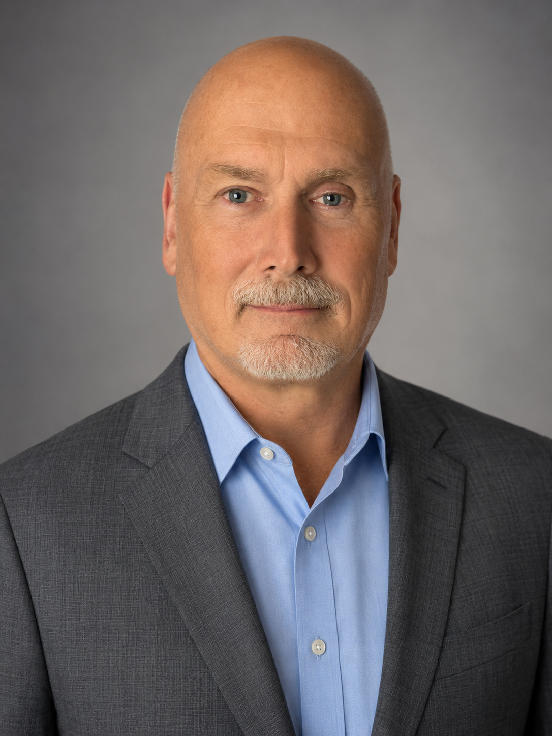 A middle-aged man with a bald head and gray beard wearing a dark gray suit and light blue dress shirt against a gray background.