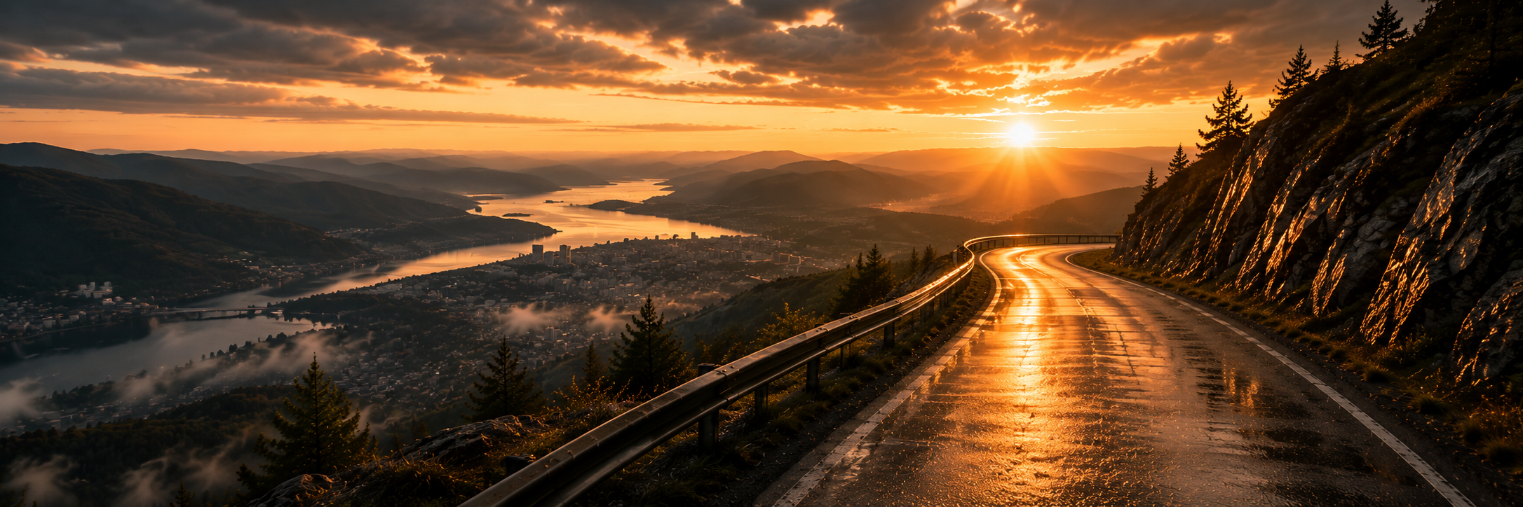 Scenic view of a winding mountain road at sunset with a reflective wet surface, overlooking a city and river valley below, surrounded by mountains and pine trees.
